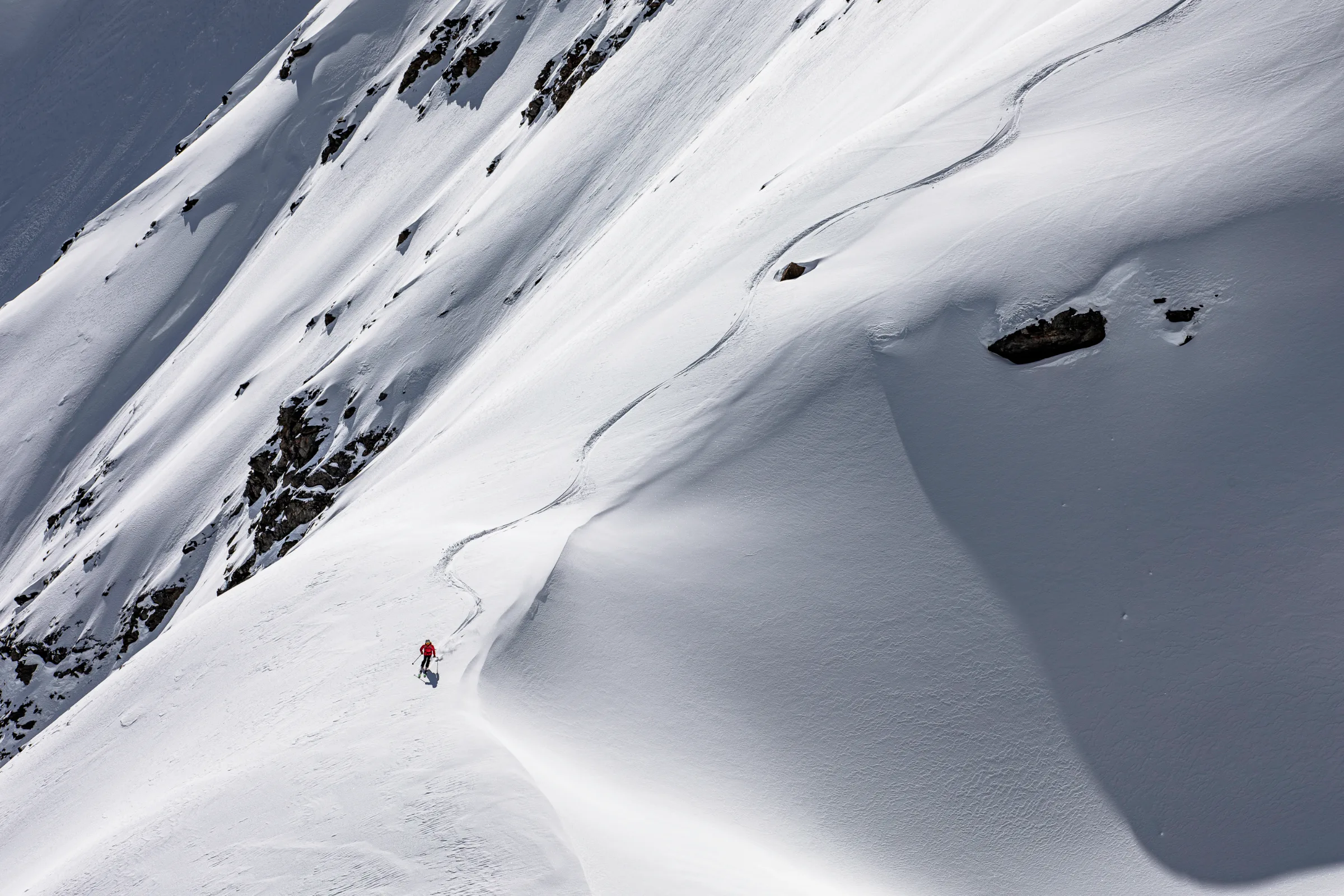 Pia Vogt skis downhill beside a large shaded snow mound, leaving a long S-shaped trail between scattered rocks behind her.