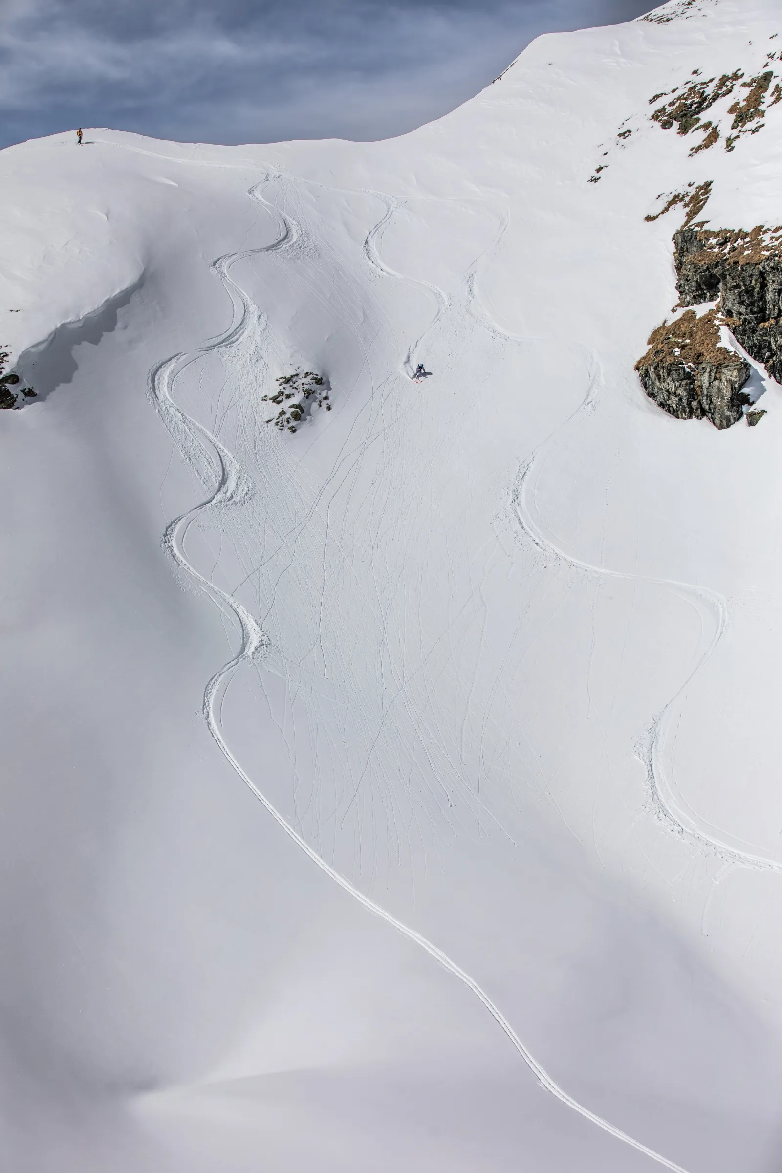 Pia Vogt skis down a broad, rock-lined snow slope in Lenzerheide while another person stands on the ridge above the curving tracks.