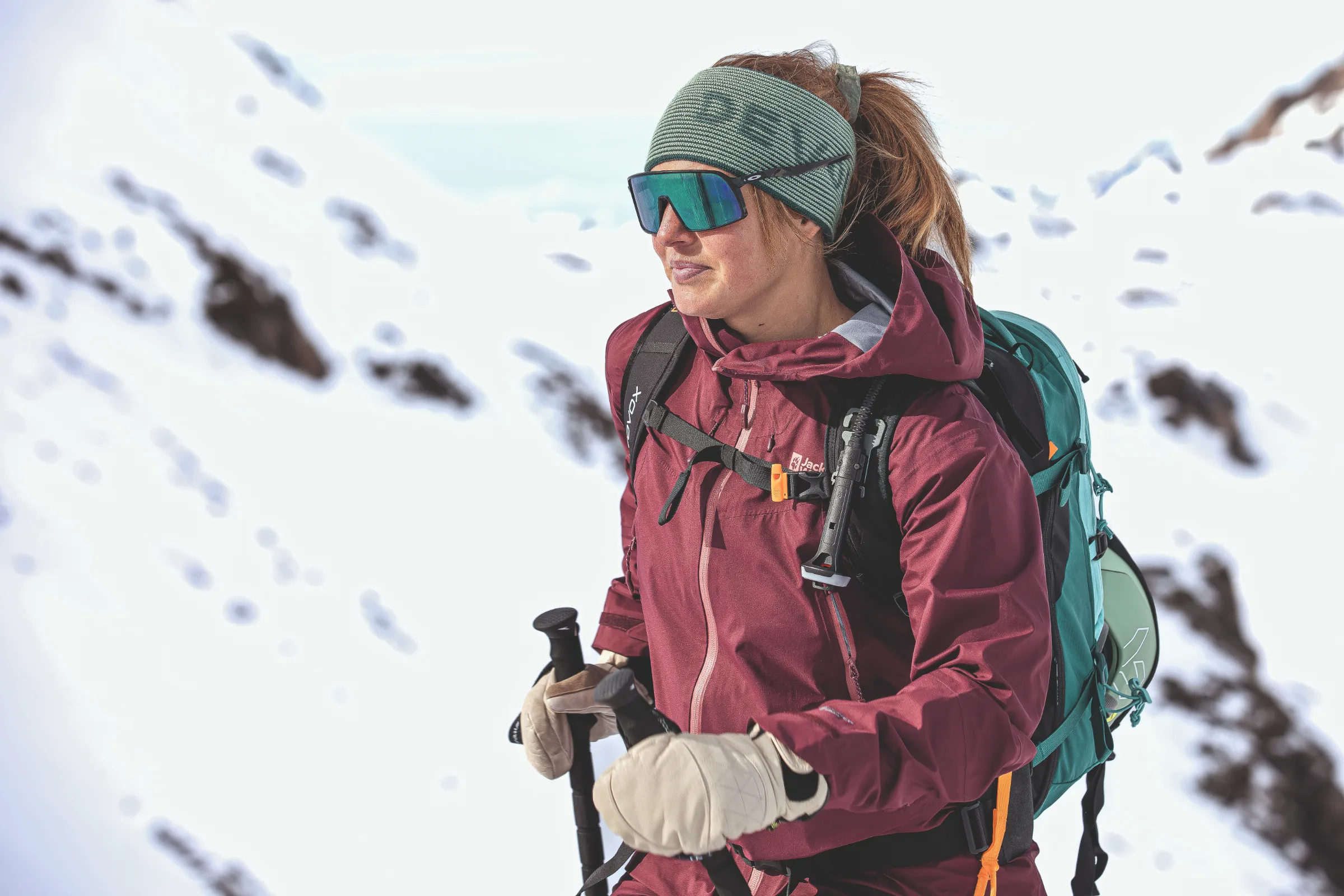 Ramona Arendt holds ski poles before a snowy rocky slope and looks to the side, wearing a green headband, mirrored sunglasses and a turquoise backpack.
