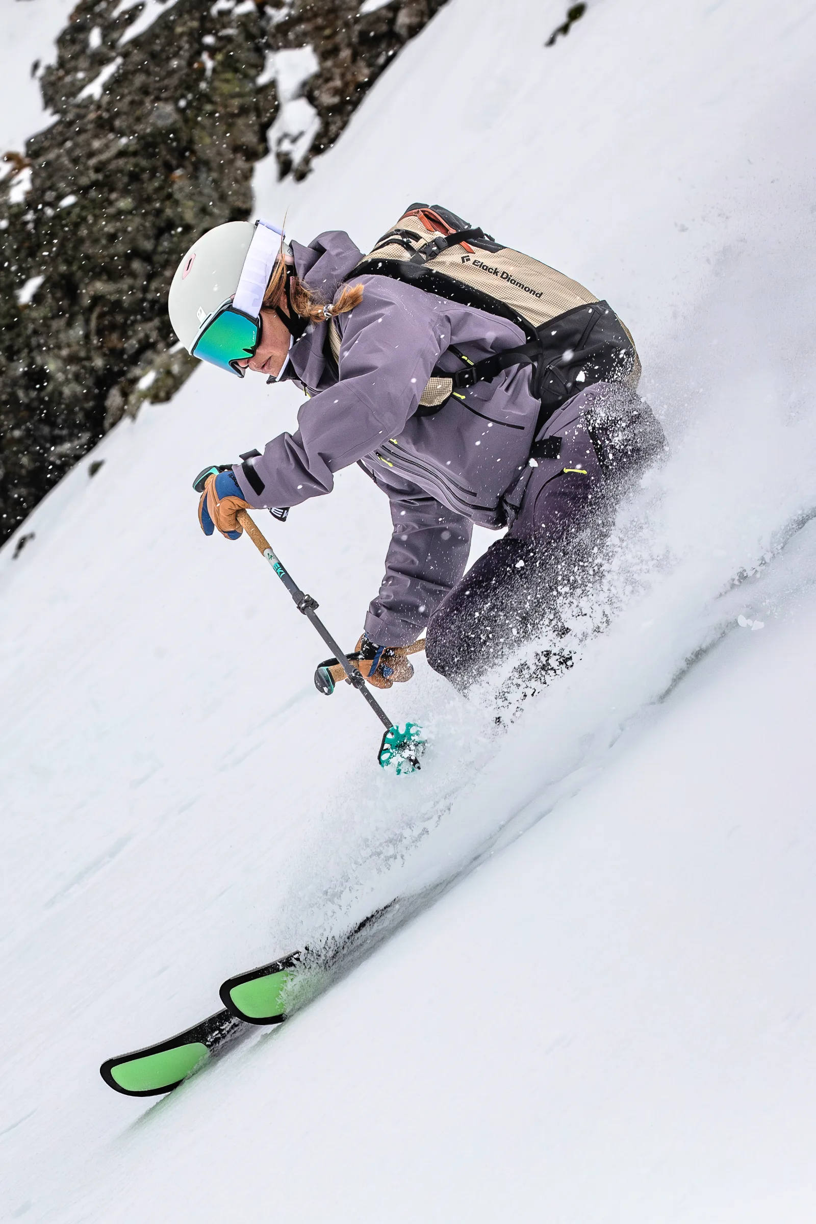 Ramona Arendt leans low into a ski turn beside a rock face, with green-and-black ski tips and flying snow in the foreground.