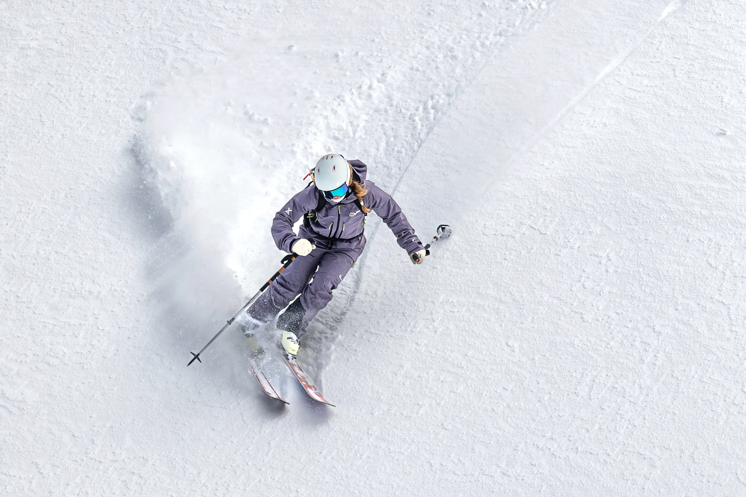 Viewed from above, Ramona Arendt turns on reddish skis across a snowy slope, leaving a curved track and a plume of snow.