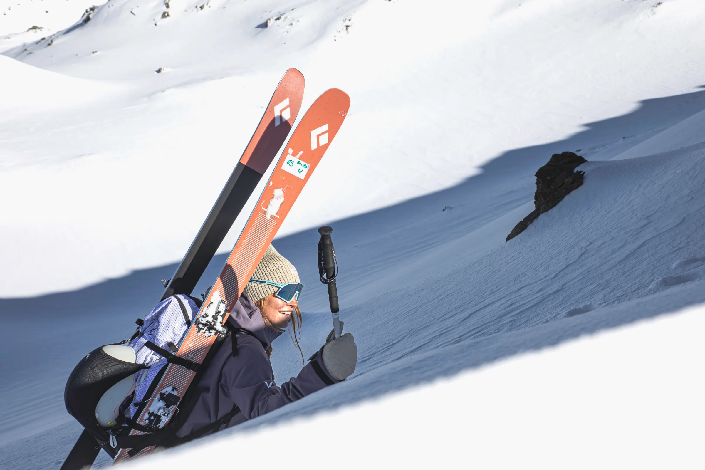 Ramona Arendt smiles as she climbs a snowy slope with red skis and a helmet on her backpack and a pole in hand, with a rock protruding from the snow behind her.
