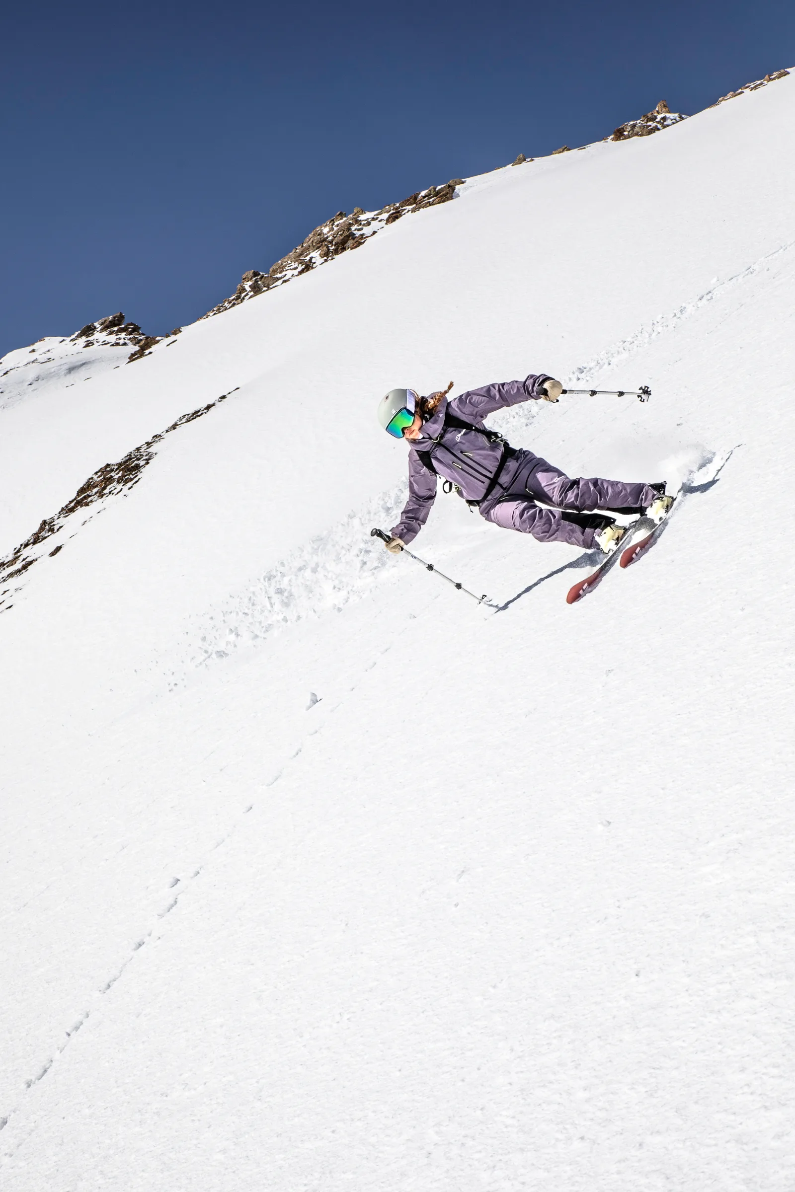 Ramona Arendt leans into a ski turn in purple outerwear, a helmet and goggles on a steep snowy slope below a rocky ridge and blue sky.