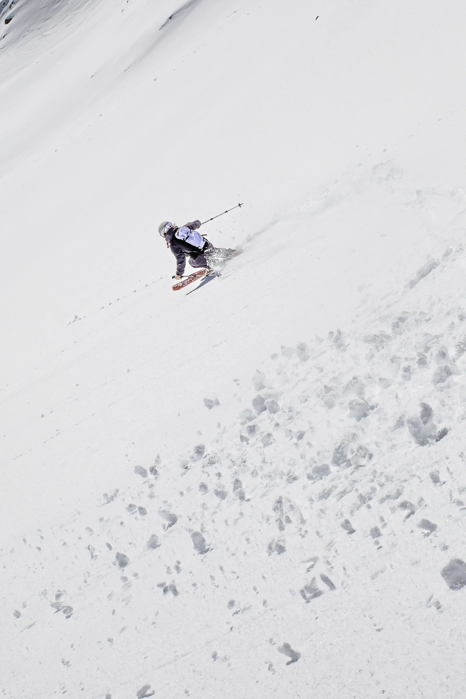 Seen from behind, Ramona Arendt skis down a broad snowy slope in a low stance with a pale backpack, throwing up snow as she turns.