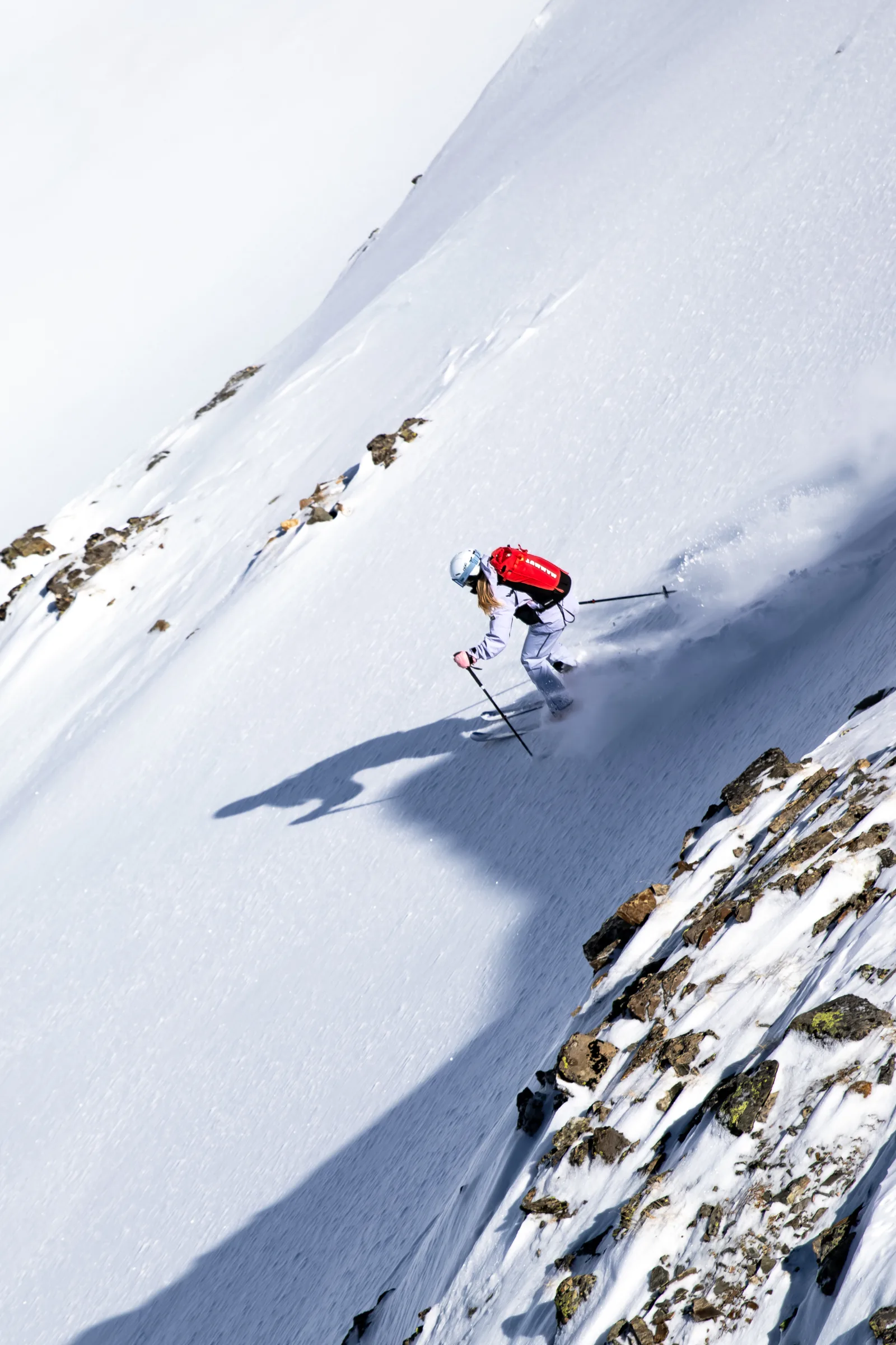 Wearing a pale ski outfit and red backpack, Ramona Arendt skis beside a rocky section of slope as snow sprays behind her skis.