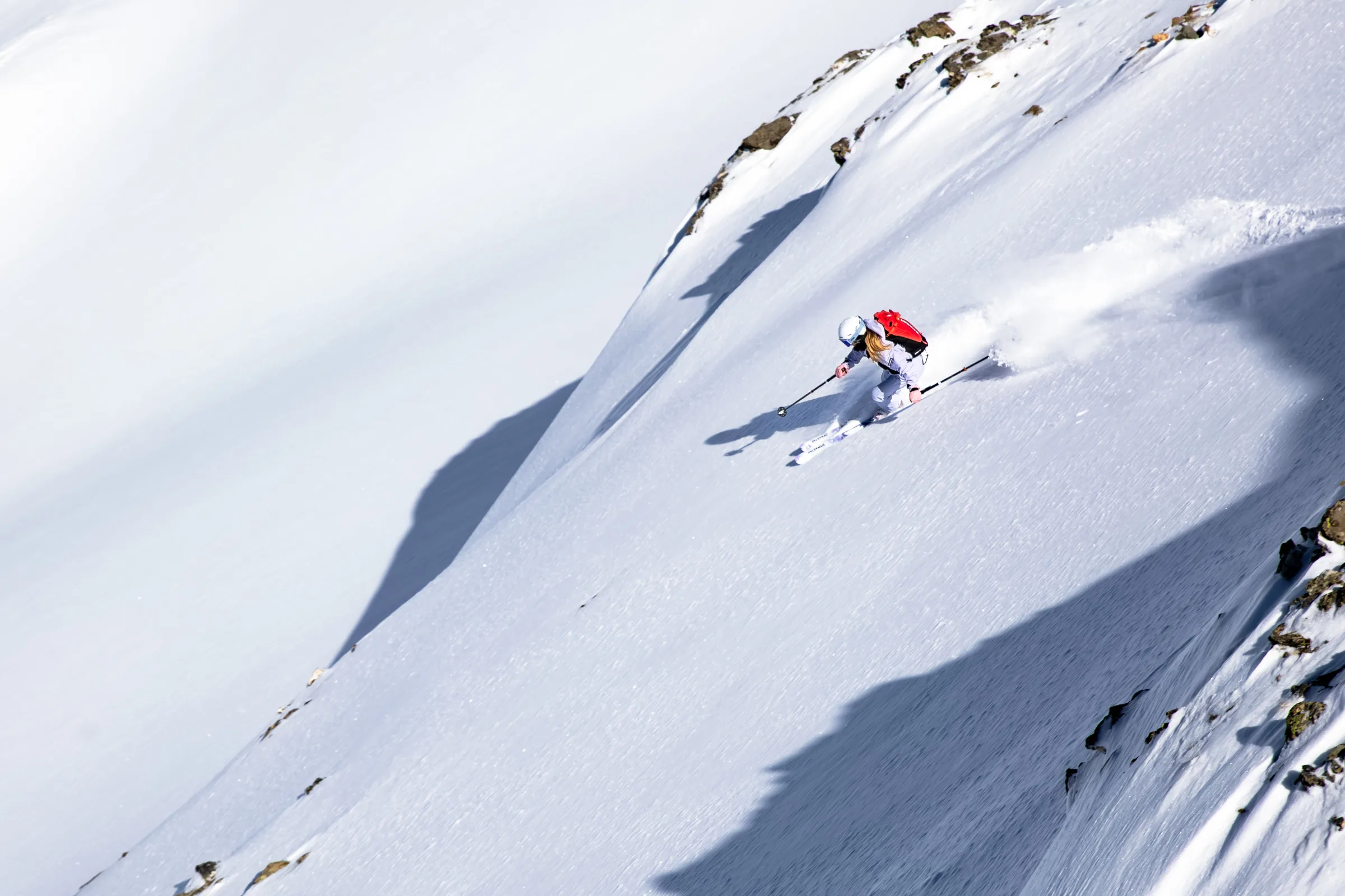 Ramona Arendt leans into a turn on a steep, sunlit snow slope, throwing up snow below exposed rocks.