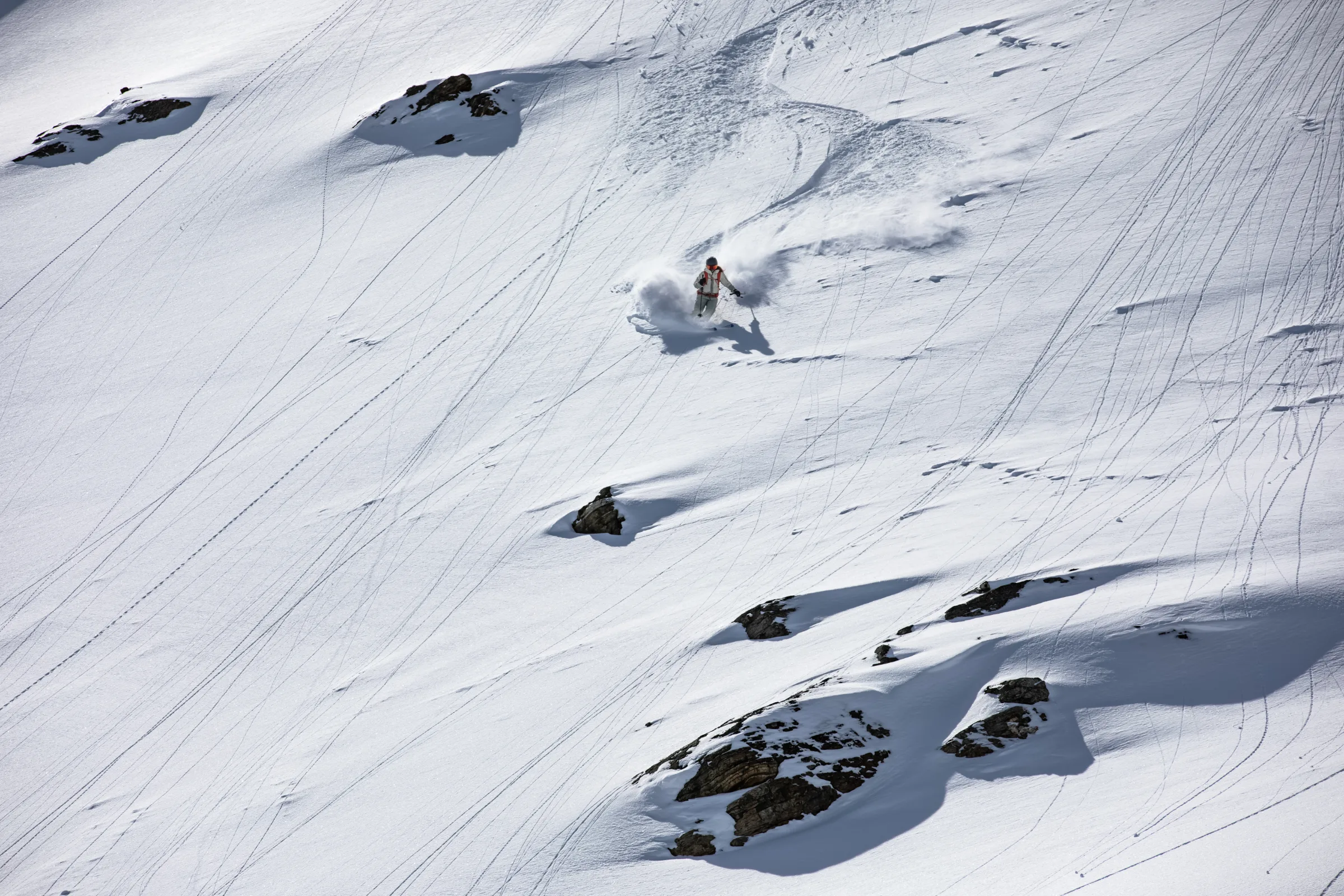 A skier turns between rocks on a broad snowy slope crisscrossed by fine tracks, leaving a wide plume of snow behind.