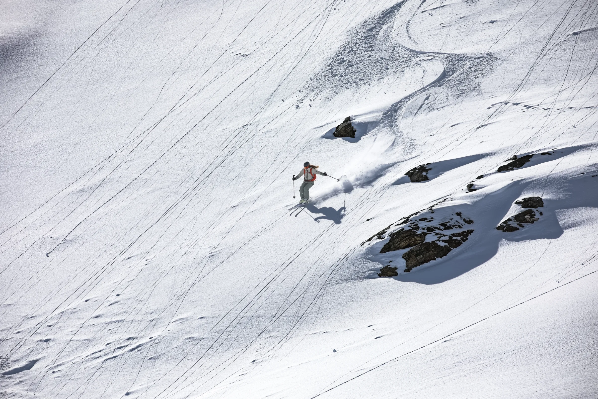 A skier descends a broad snowy slope to the left of snow-covered rocks, leaving a plume of snow below her curving track.