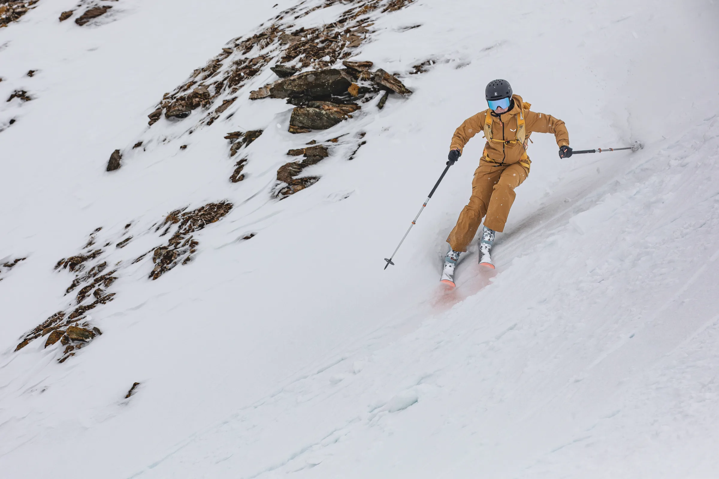 Sabine Speidel skis down a rocky snow slope in Lenzerheide, wearing ochre clothing and holding her poles wide.