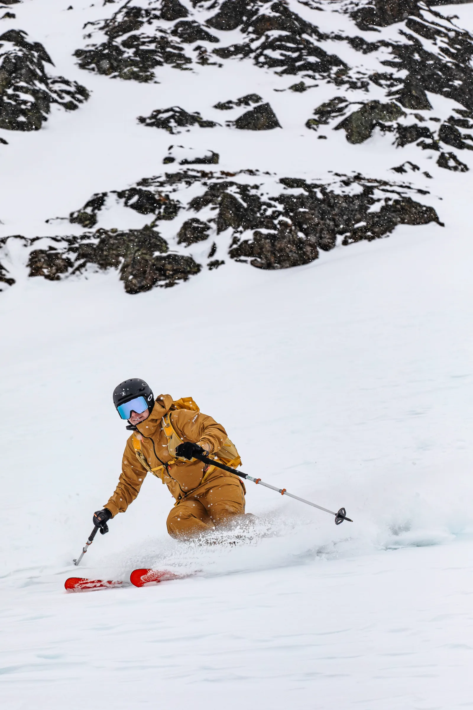 Sabine Speidel leans low into a turn on a snowy slope beneath dark rocks, with snow spraying around her knees.