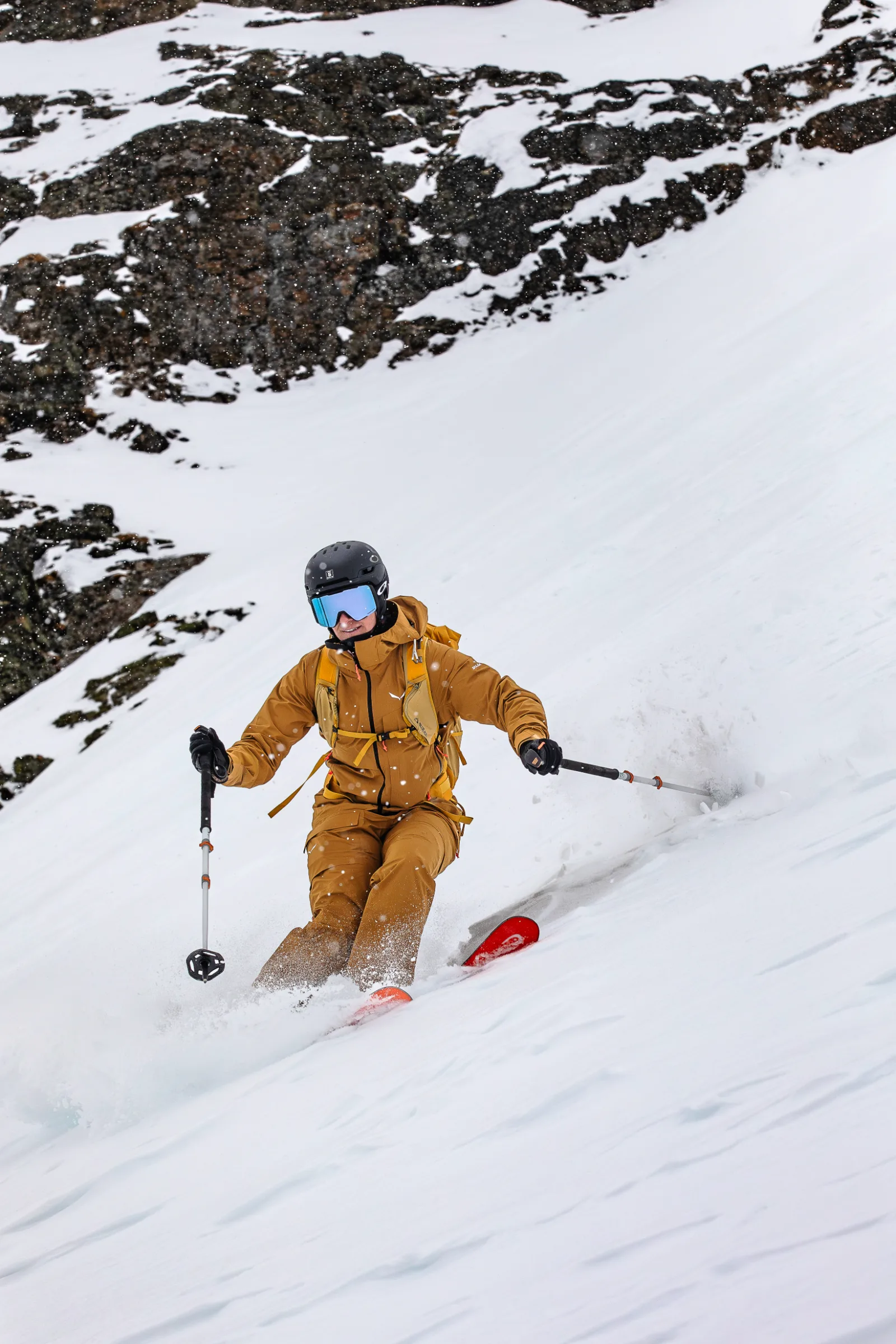 Wearing a black helmet and blue goggles, Sabine Speidel skis through spraying snow down a steep slope below snow-covered rocks.