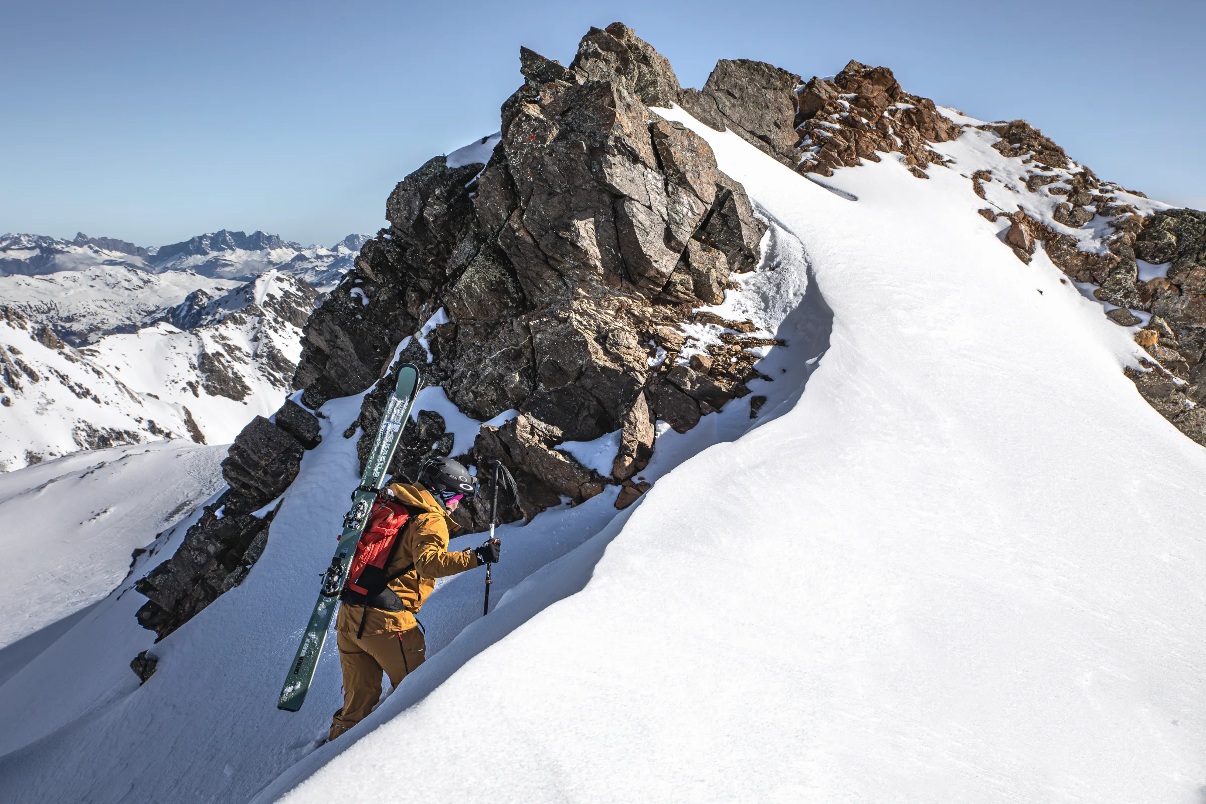 Sabine Speidel climbs a narrow snow gully beside a rocky ridge with skis on her red backpack, with snowy mountains stretching behind her.