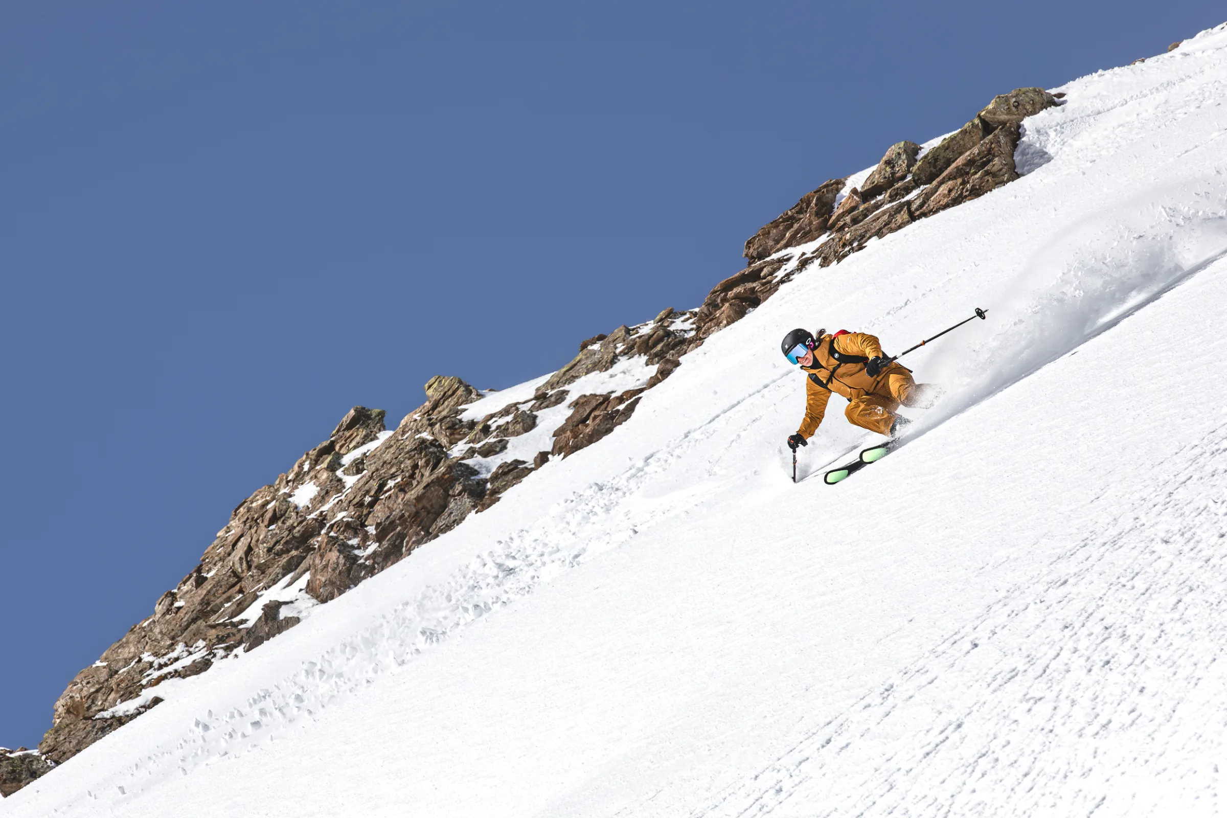 Sabine Speidel makes a low turn on a steep snow slope beside rocks under a blue sky, trailing a plume of snow.