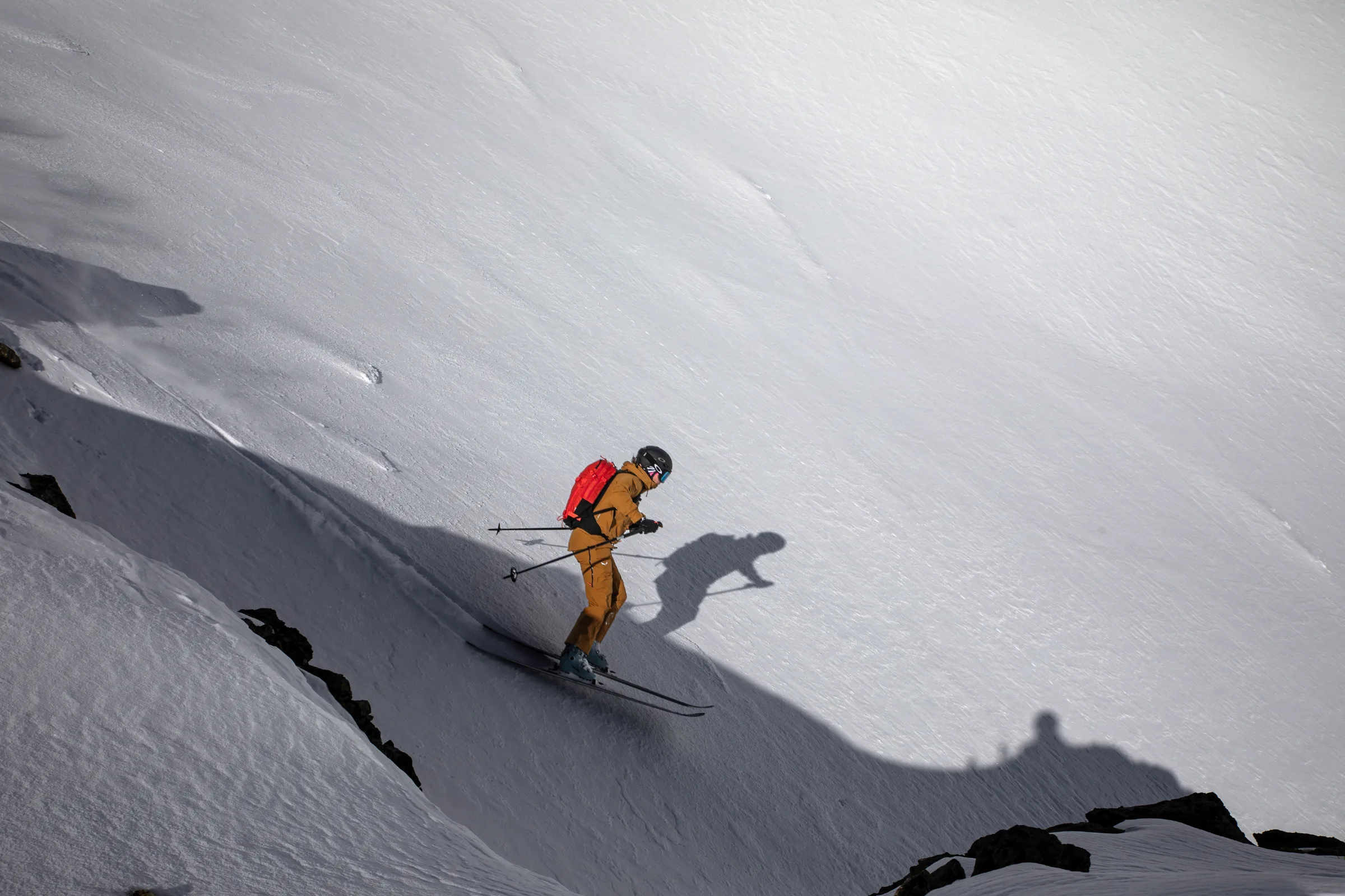 Sabine Speidel skis through a shaded snow hollow between rocks with a red backpack, casting a distinct shadow onto the bright slope beside her.