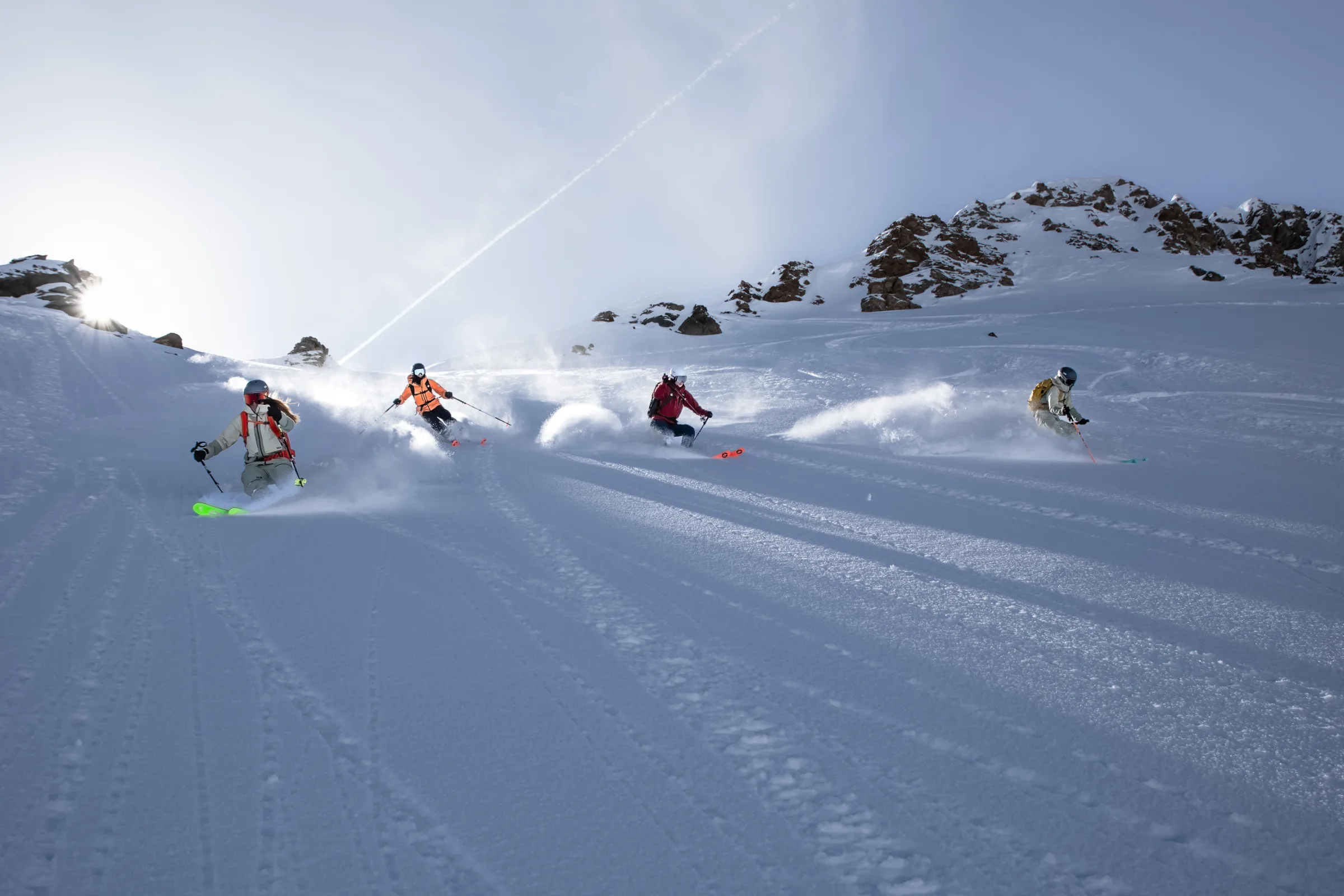 Vier Personen ziehen auf Ski Schwünge durch den Pulverschnee unter einem felsigen Grat in Lenzerheide und wirbeln Schneefahnen auf.