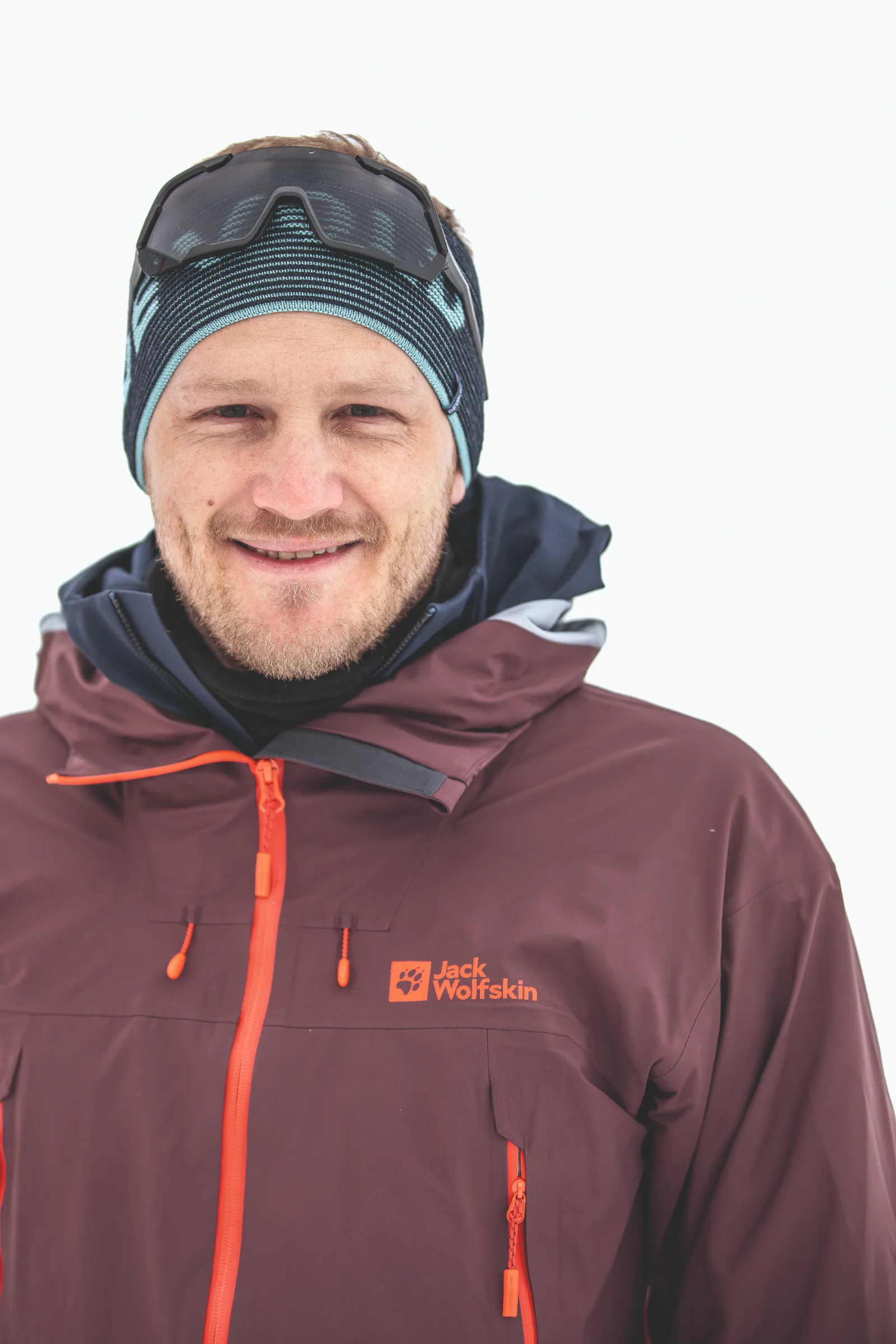 Simon Stickl smiles at the camera against a white background, wearing a striped headband, sunglasses pushed up on his head and a burgundy jacket.