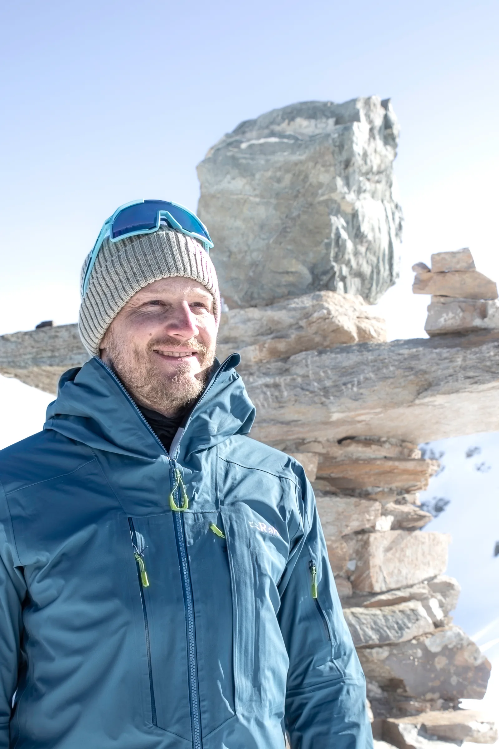 Simon Stickl smiles in a blue jacket and grey knit hat with sunglasses perched on top, beside stacked stone slabs in the snow.