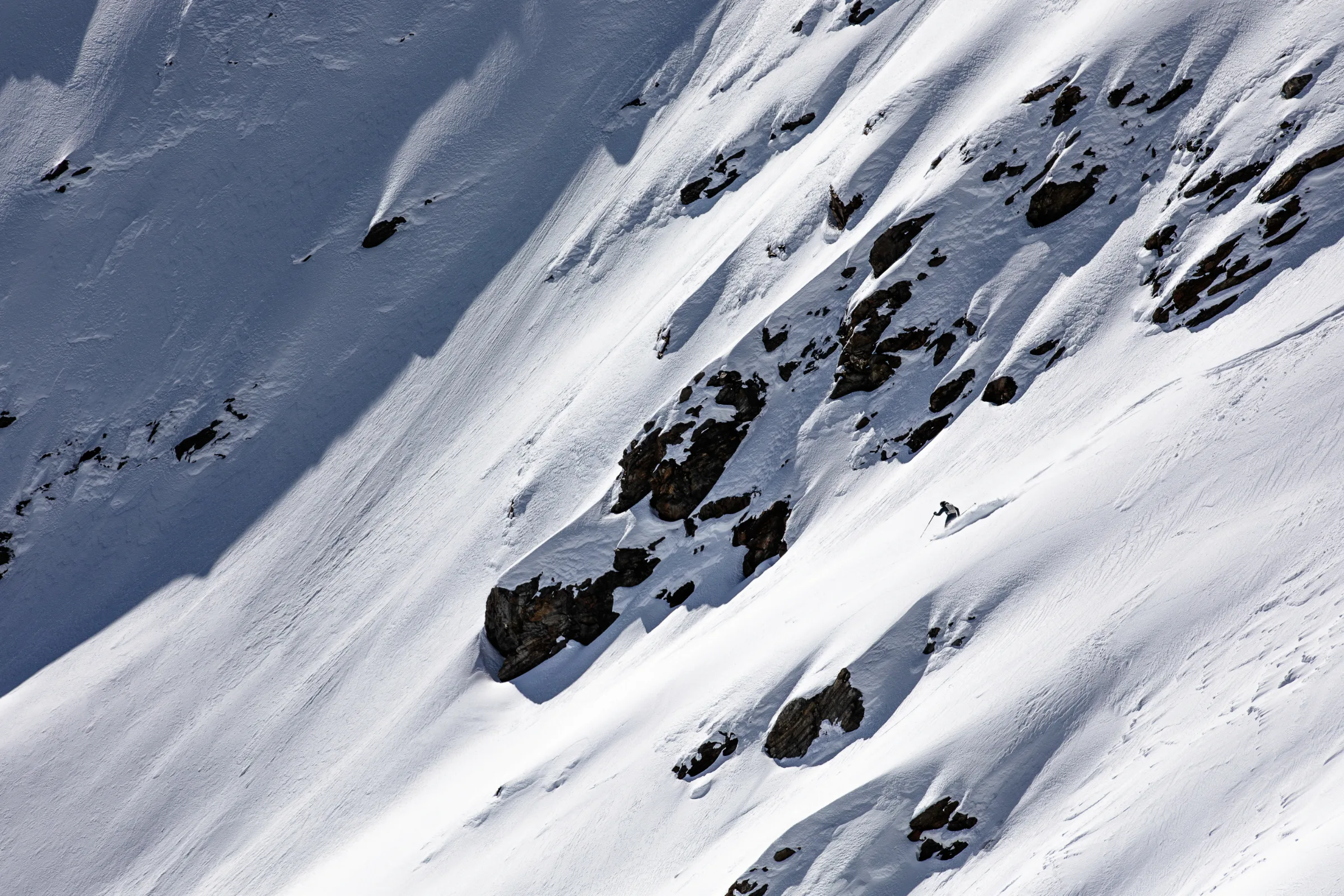 Simon Stickl skis through snow to the right of a dark rock band, leaving a curved track across the steep slope.