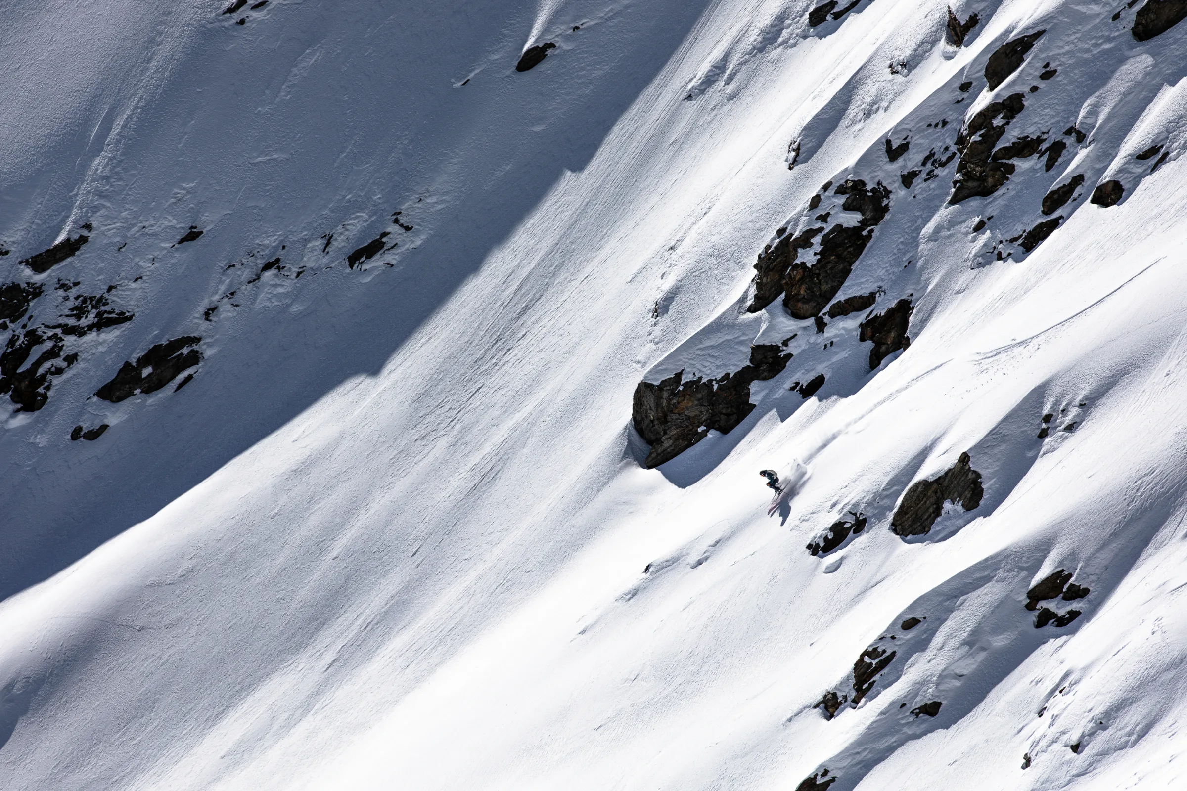 Simon Stickl skis down a steep snowy slope between rocks with knees bent, spraying snow behind his skis.