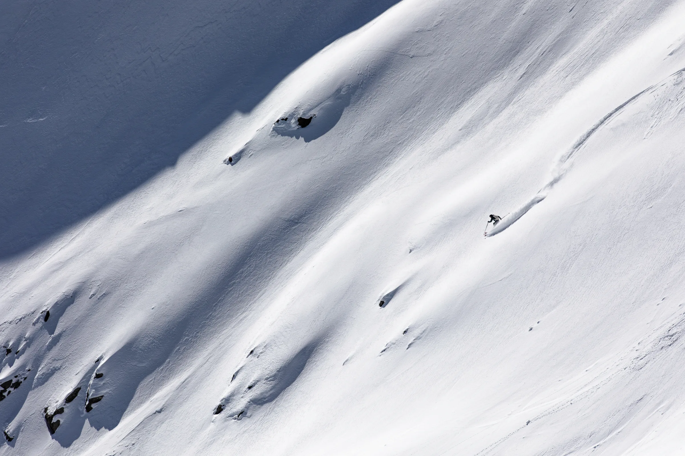 Simon Stickl skis across a broad snowy slope dotted with rocks, his long track trailing up to the right.
