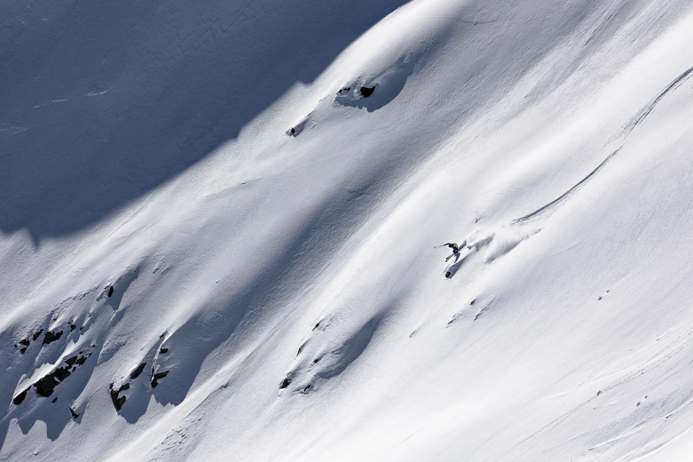 Simon Stickl leans into a turn beside a small rock, throwing a broad fan of snow across the steep slope.