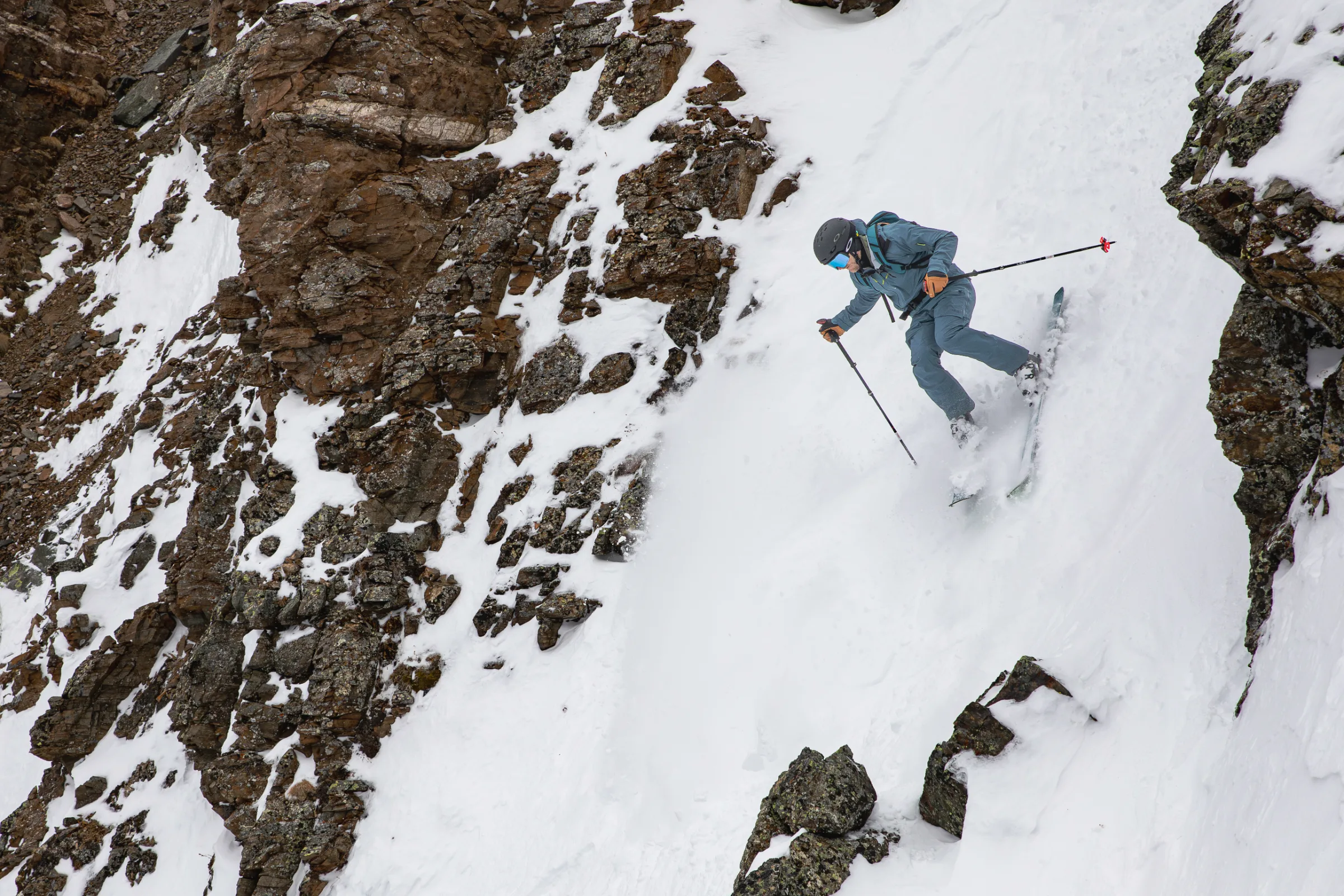 Simon Stickl skis through a narrow snowy chute between brown rock walls, wearing blue ski clothing, a helmet, goggles and a backpack.