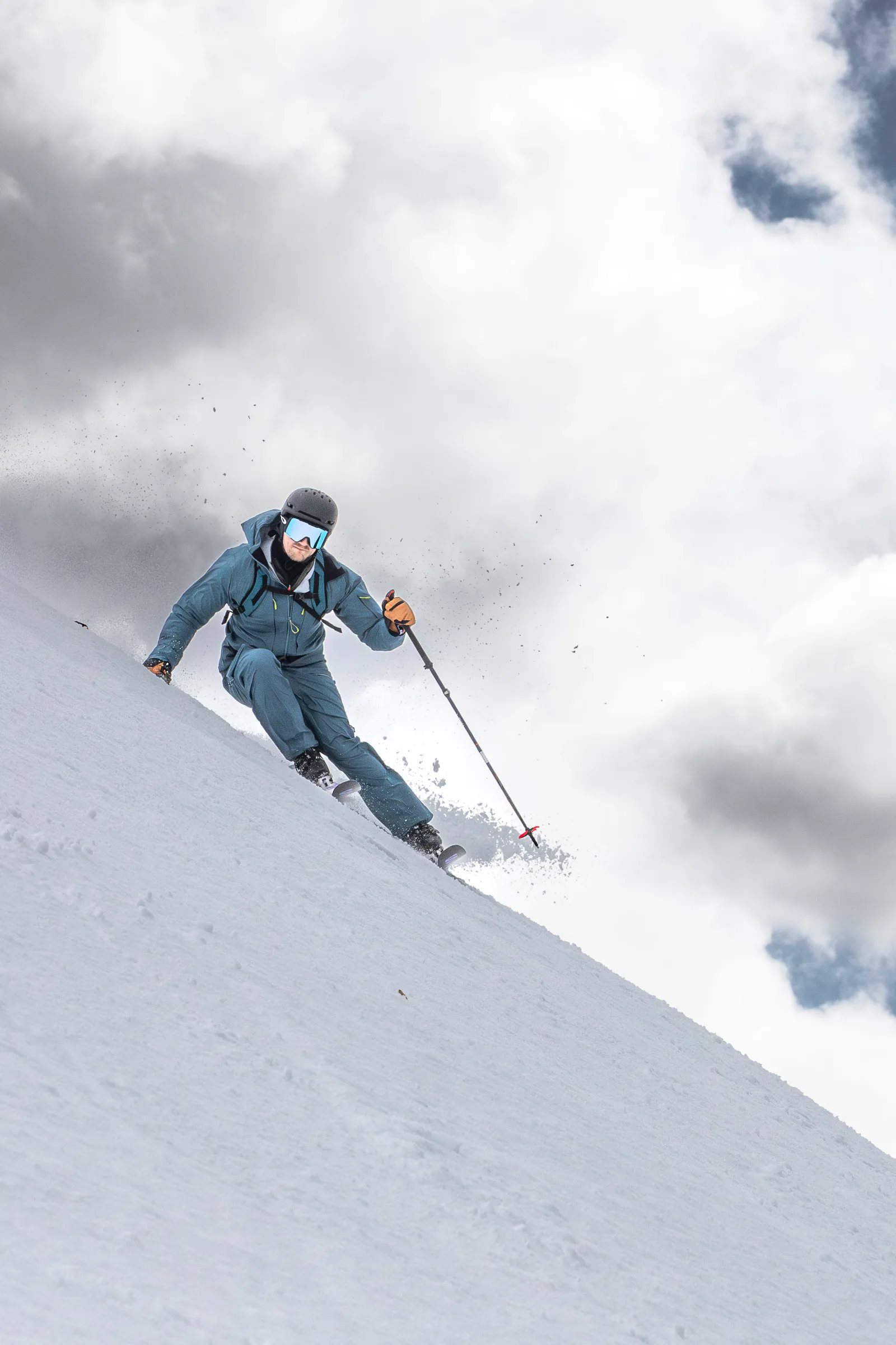 Seen from below, Simon Stickl skis down a snowy slope with bent knees, spraying snow against a cloudy sky.
