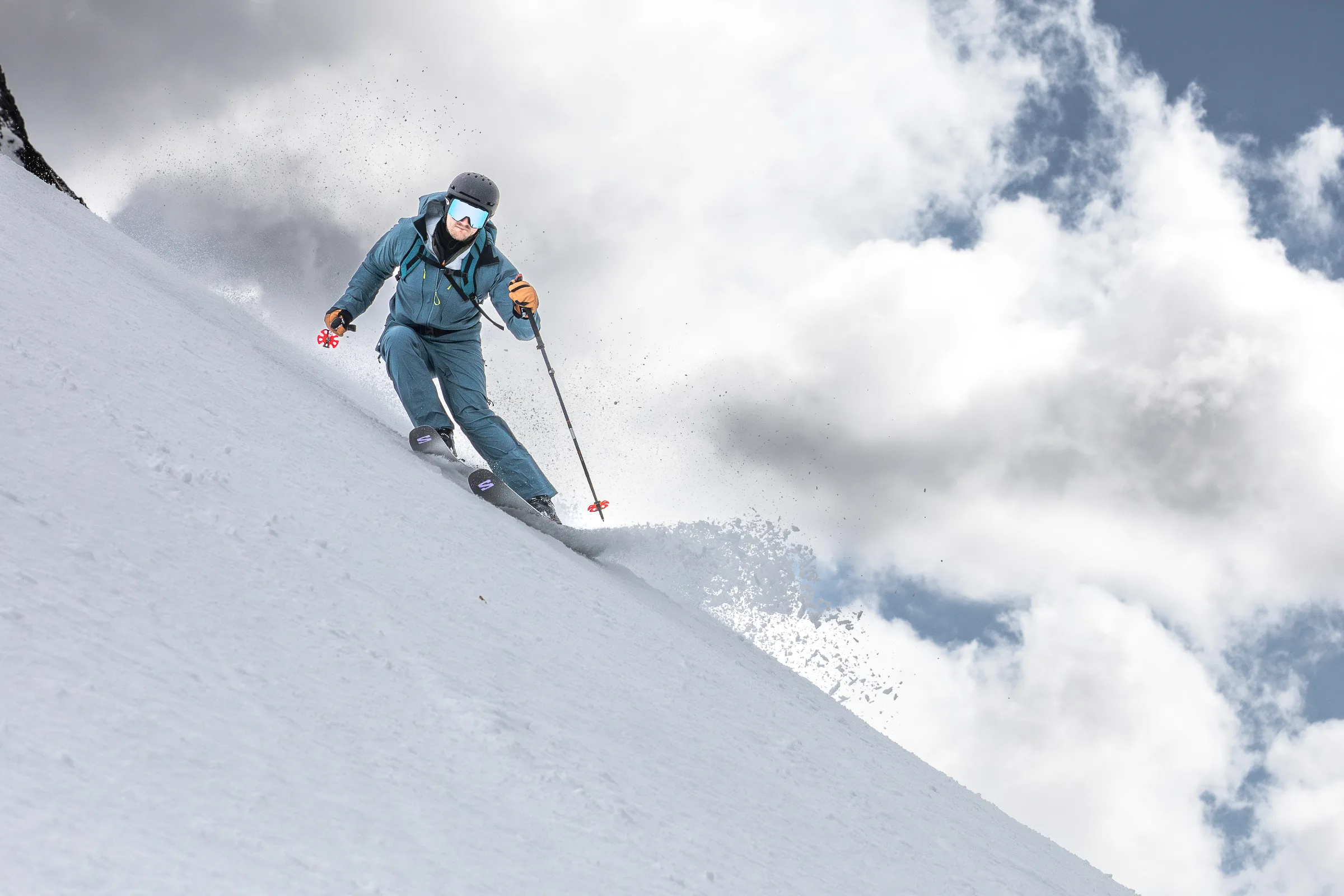 Simon Stickl edges his skis on a steep snowy slope with his poles held out to the sides, sending snow flying against a cloudy sky.