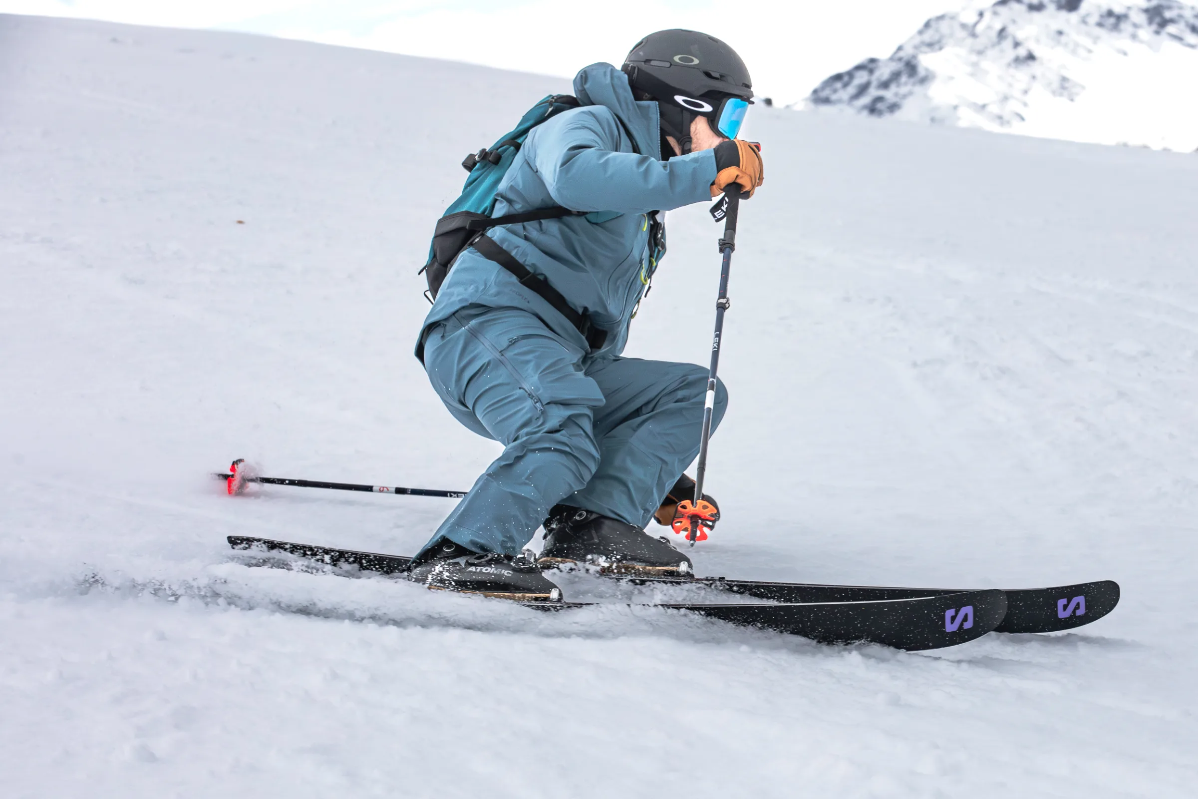 Simon Stickl makes a low, bent-knee turn on a snowy slope, holding ski poles, with snow-covered rocks in the background.
