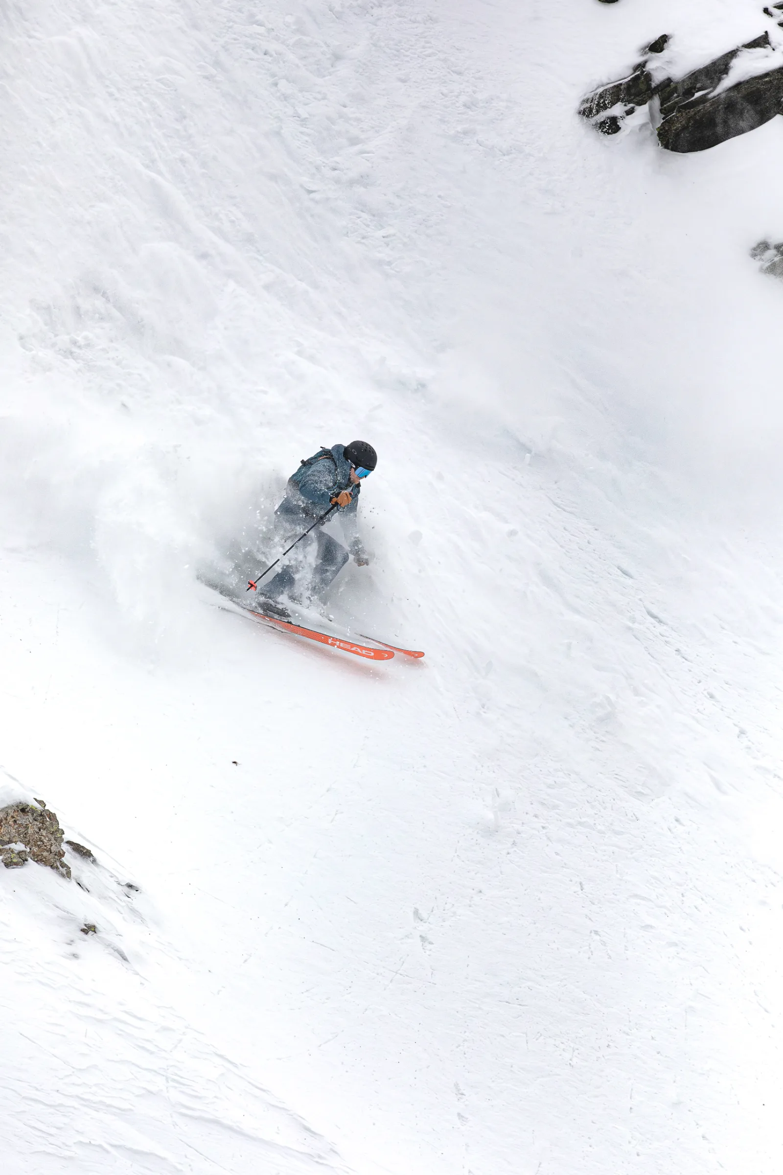 Simon Stickl skis on orange skis through a steep, rocky snow gully in Lenzerheide, throwing up a spray of snow.