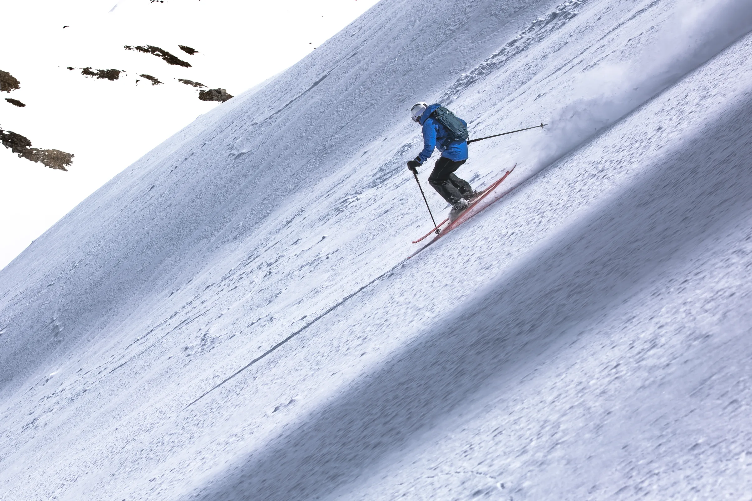 Seen from behind, Simon Stickl skis down a steep slope on red skis with a backpack, trailing a plume of snow as rocks protrude from the snow on the left.