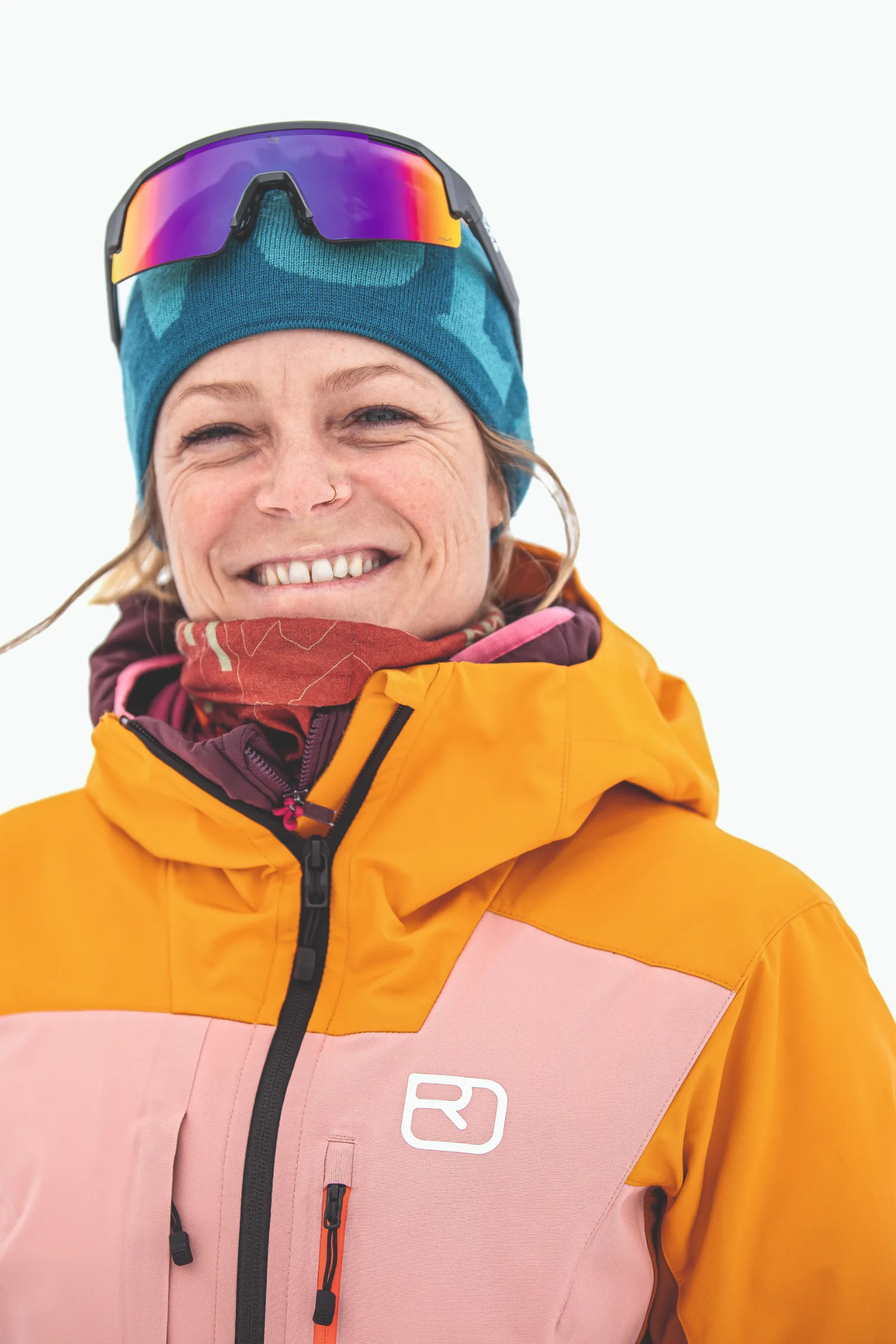 Sofia Muigg smiles against a white background, wearing a blue beanie with mirrored sports glasses perched on top and an orange-and-pink ski jacket.