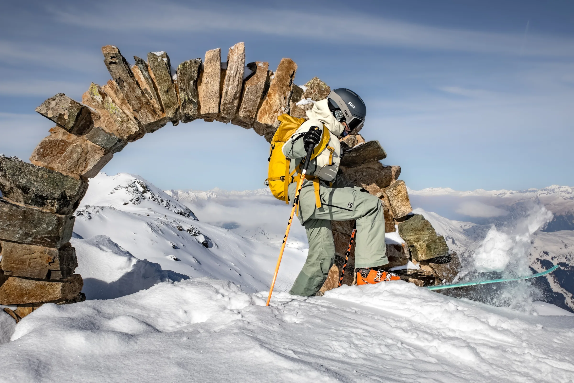 Sofia Muigg lifts a ski beside a stone arch, scattering snow, with a yellow backpack and the snowy mountains of Lenzerheide behind her.