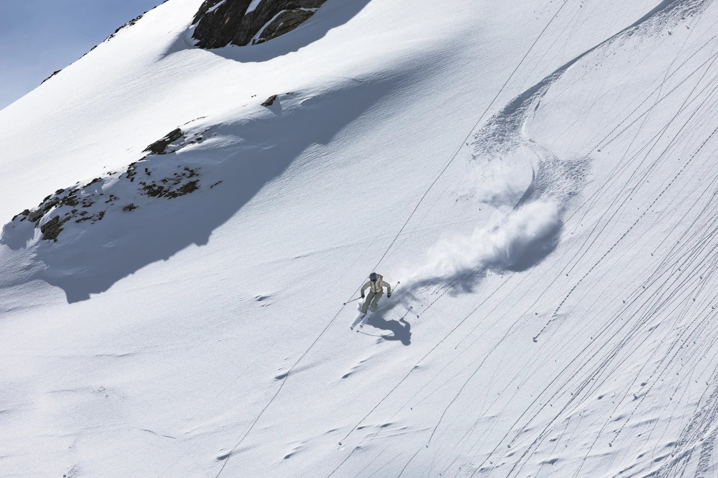Sofia Muigg skis down a sunlit snowy slope with her poles held out, leaving a cloud of snow and curved tracks below shadowed rocks.