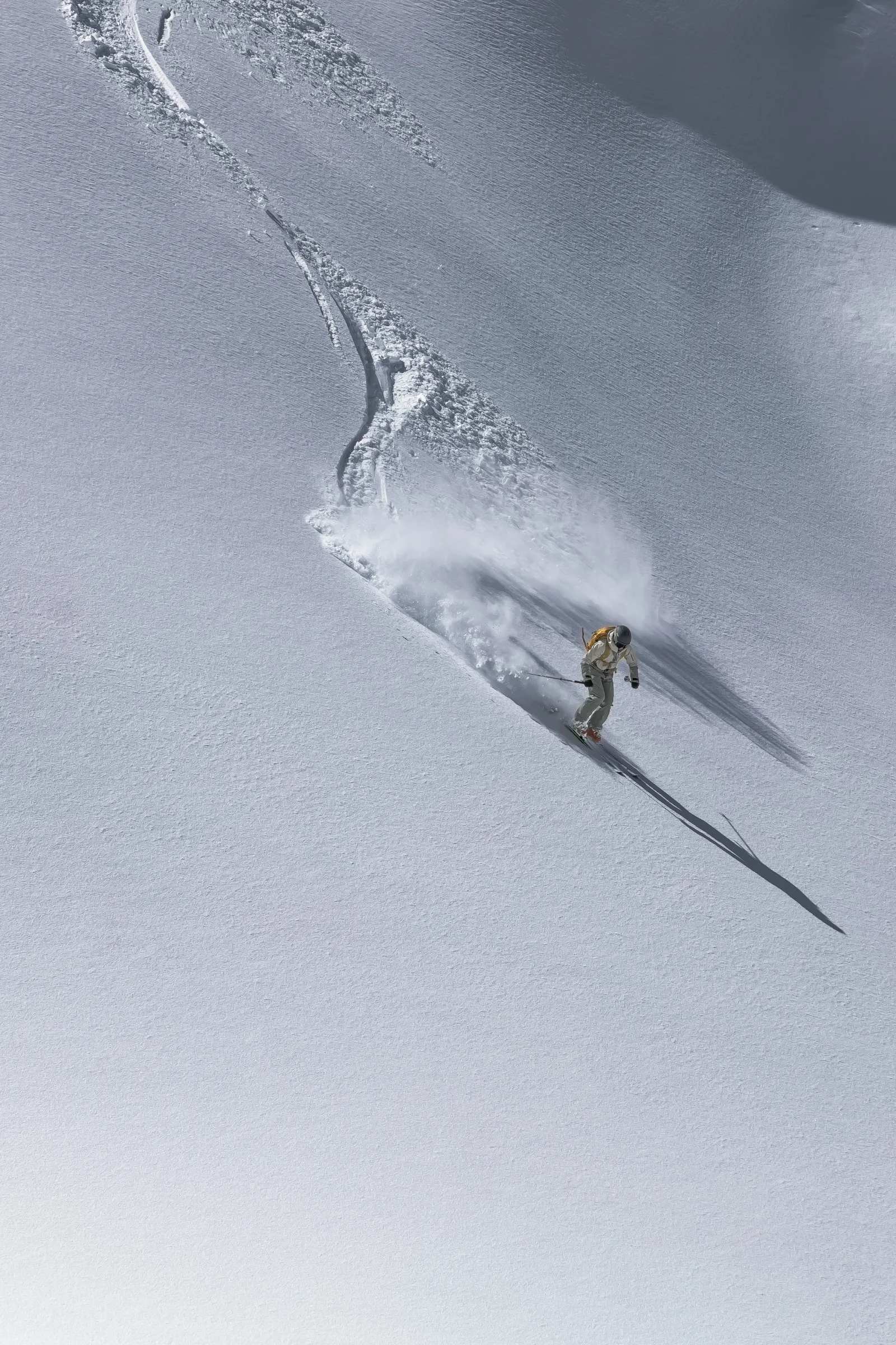 Sofia Muigg skis through an open powder slope with a yellow backpack, leaving a curving track and casting a long shadow to the right.