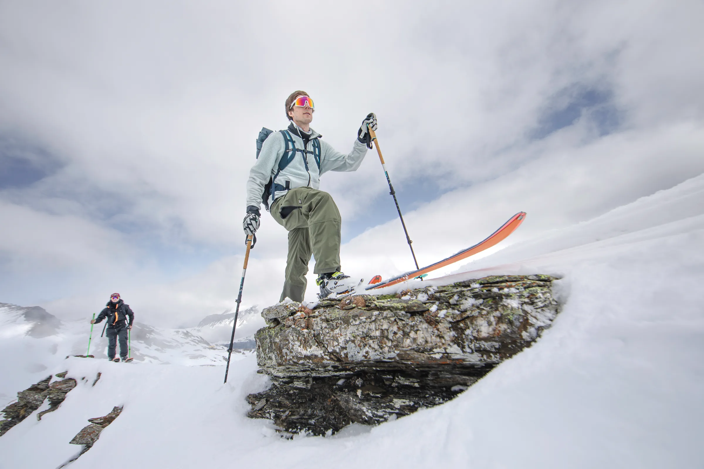 Valentin Rapp steps over a snow-covered rock ledge on touring skis with poles, while another person follows in the background.