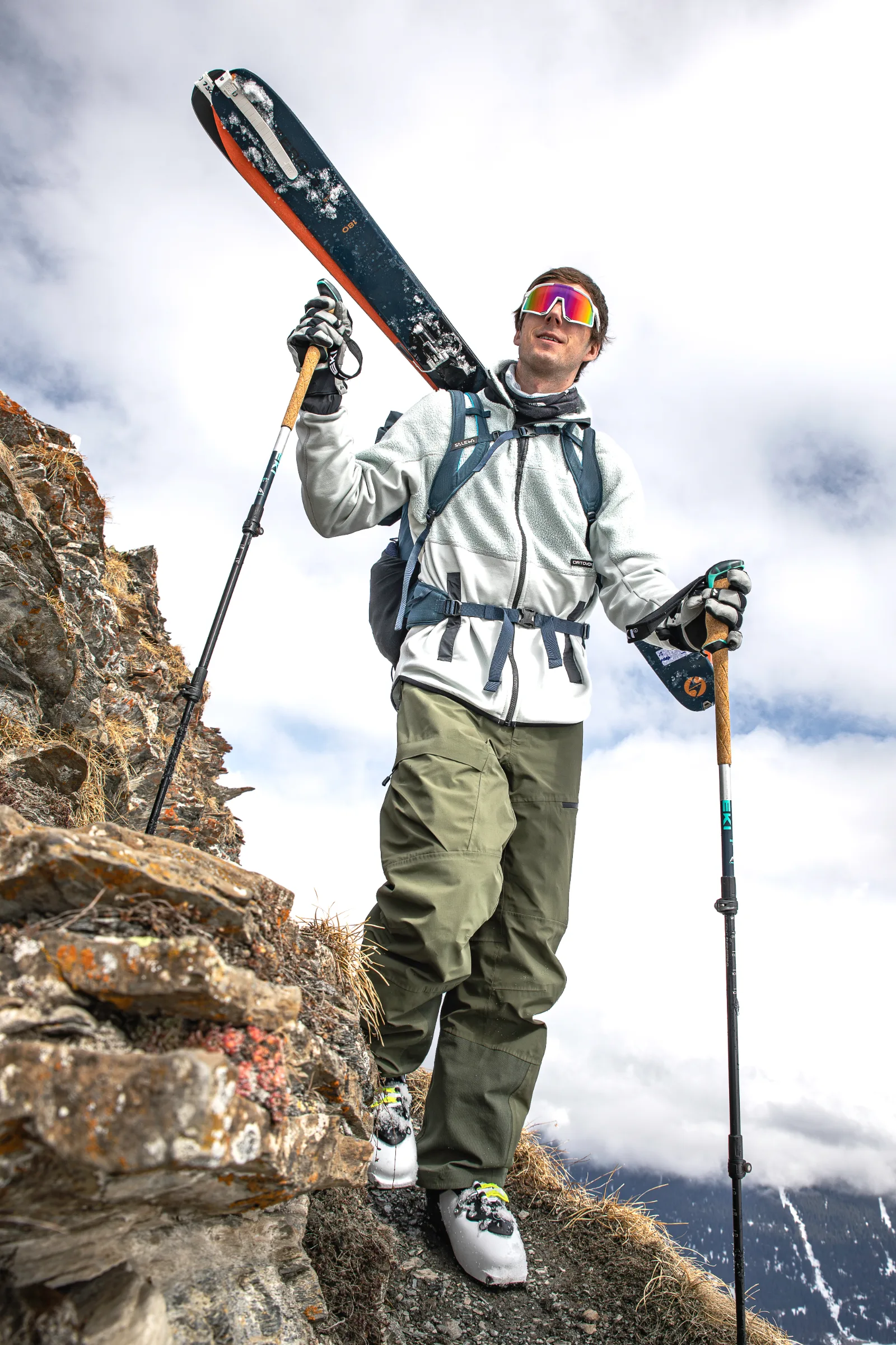 Valentin Rapp walks along a narrow rocky mountain path with skis on his backpack and poles in his hands beneath a cloudy sky.