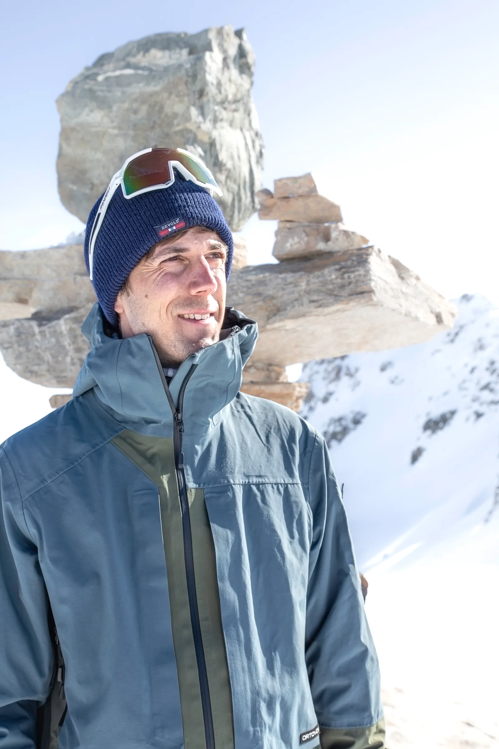 Valentin Rapp smiles and looks aside, wearing a blue beanie with sunglasses perched on top, with stacked stones and snowy mountains behind him.