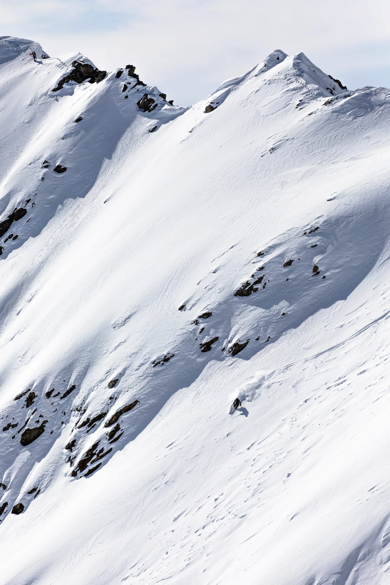 Valentin Rapp skis down a broad, rocky steep slope near Lenzerheide, trailing a plume of snow, while another person stands on the ridge above.