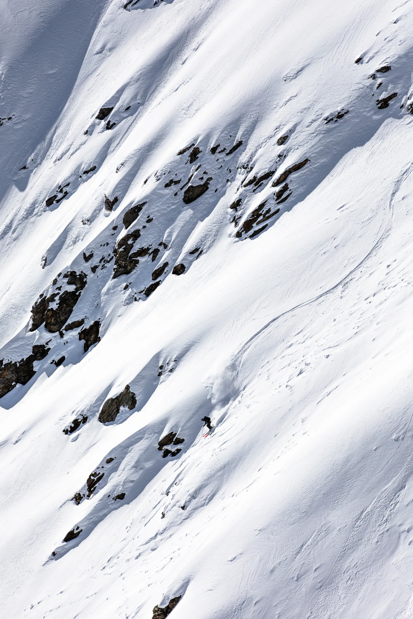 Valentin Rapp skis down a steep snowy slope beside exposed rocks, leaving a curved track and a spray of snow behind him.