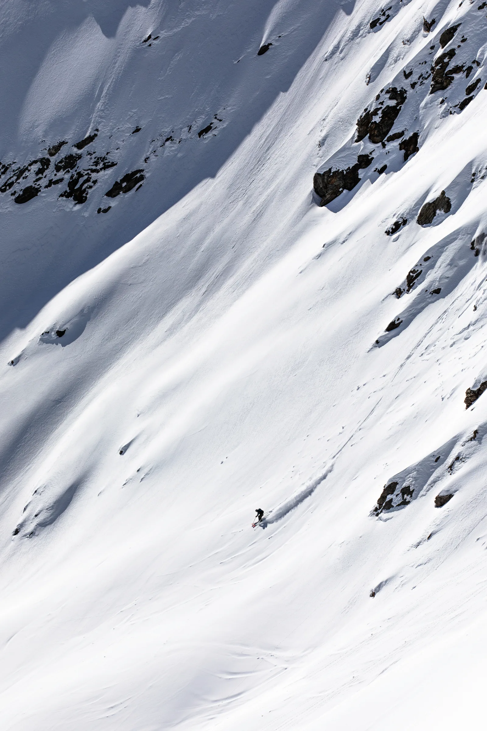 Valentin Rapp makes a turn across a broad snowy slope beneath dark rocks, throwing up a long plume of snow.