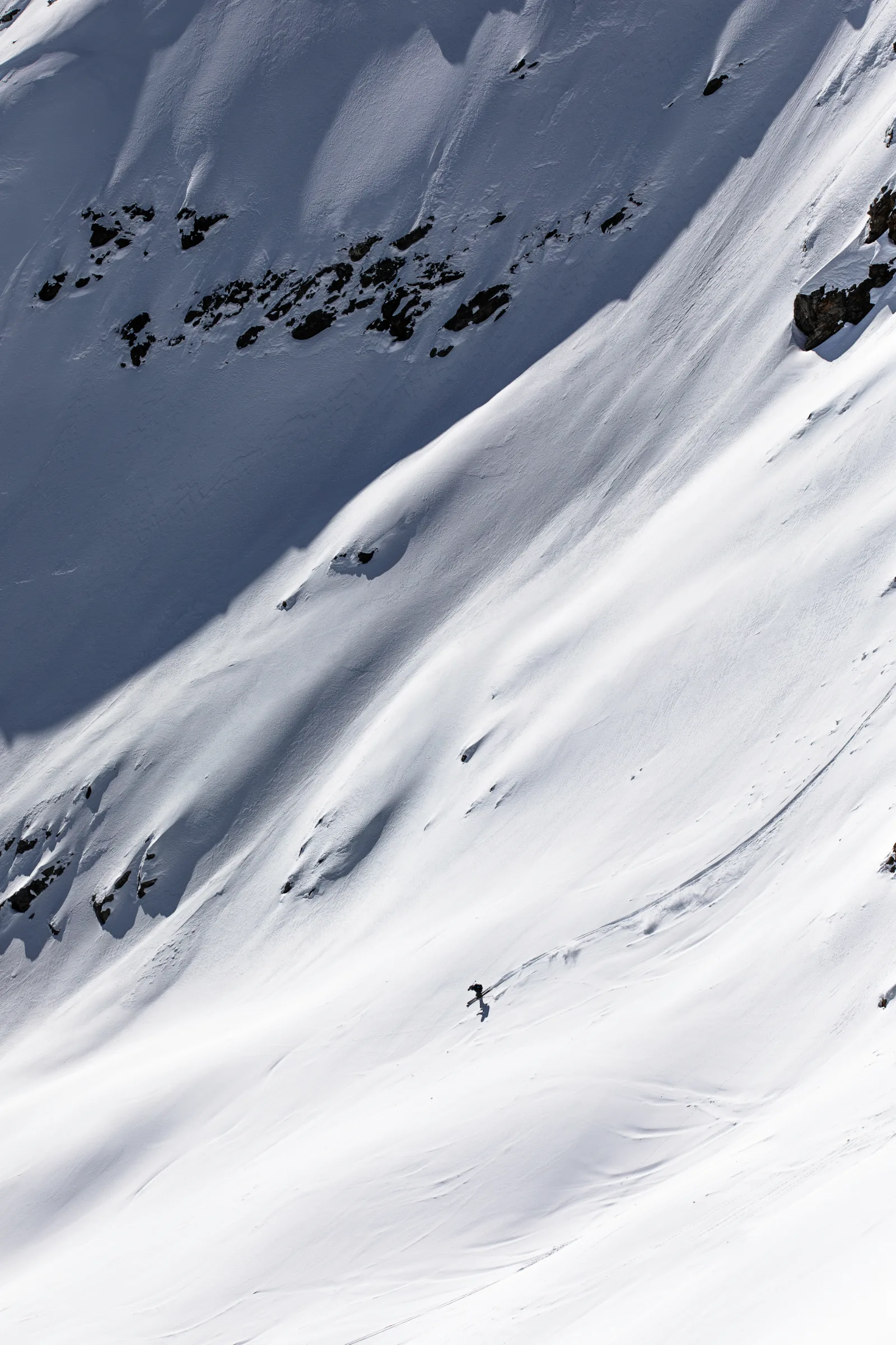 Valentin Rapp skis across an open slope patterned with sun and shadow, his long curved track extending uphill to the right.