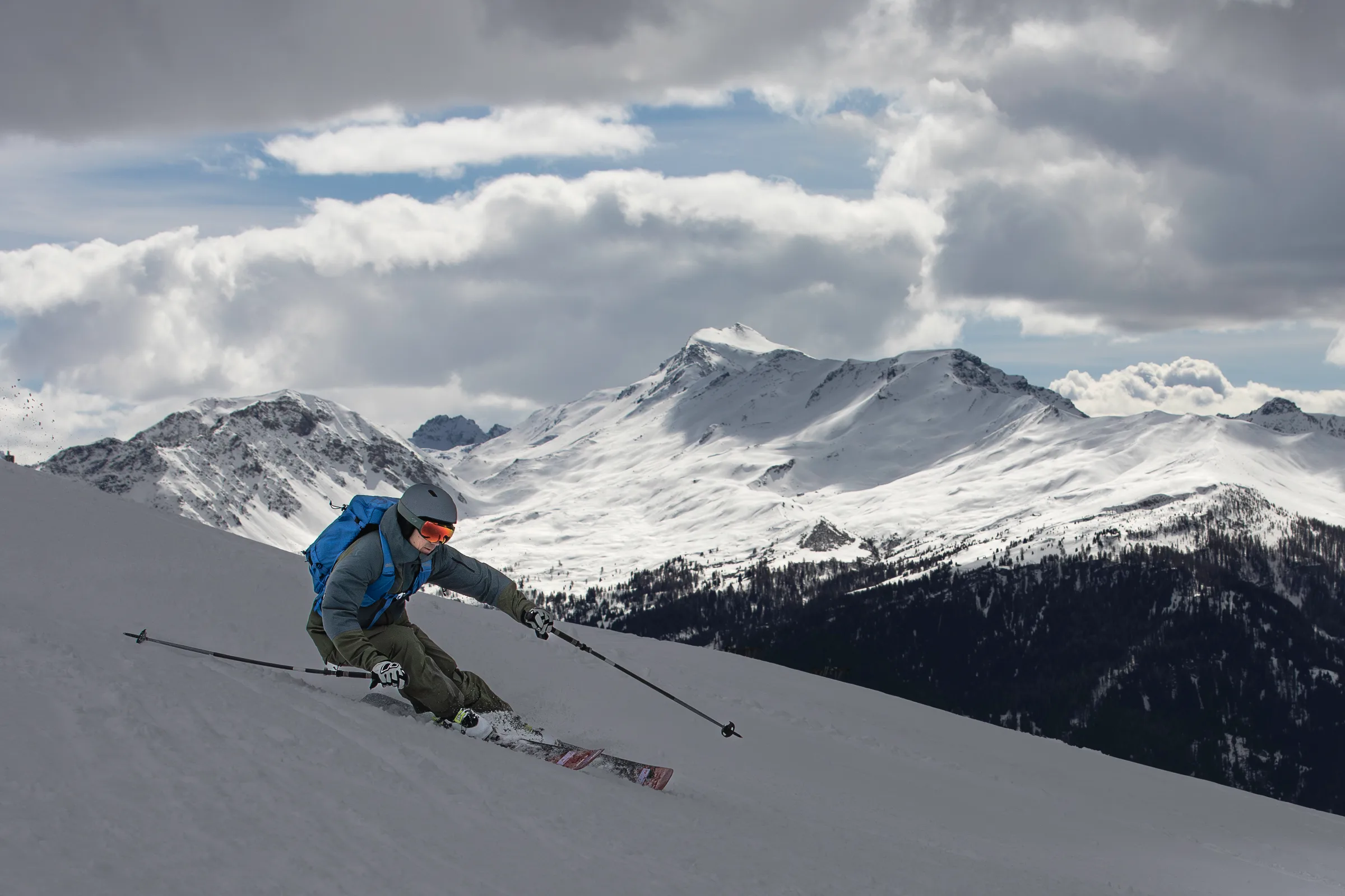 Valentin Rapp leans into a ski turn wearing a helmet and blue backpack, with forested slopes and snowy peaks near Lenzerheide behind him.