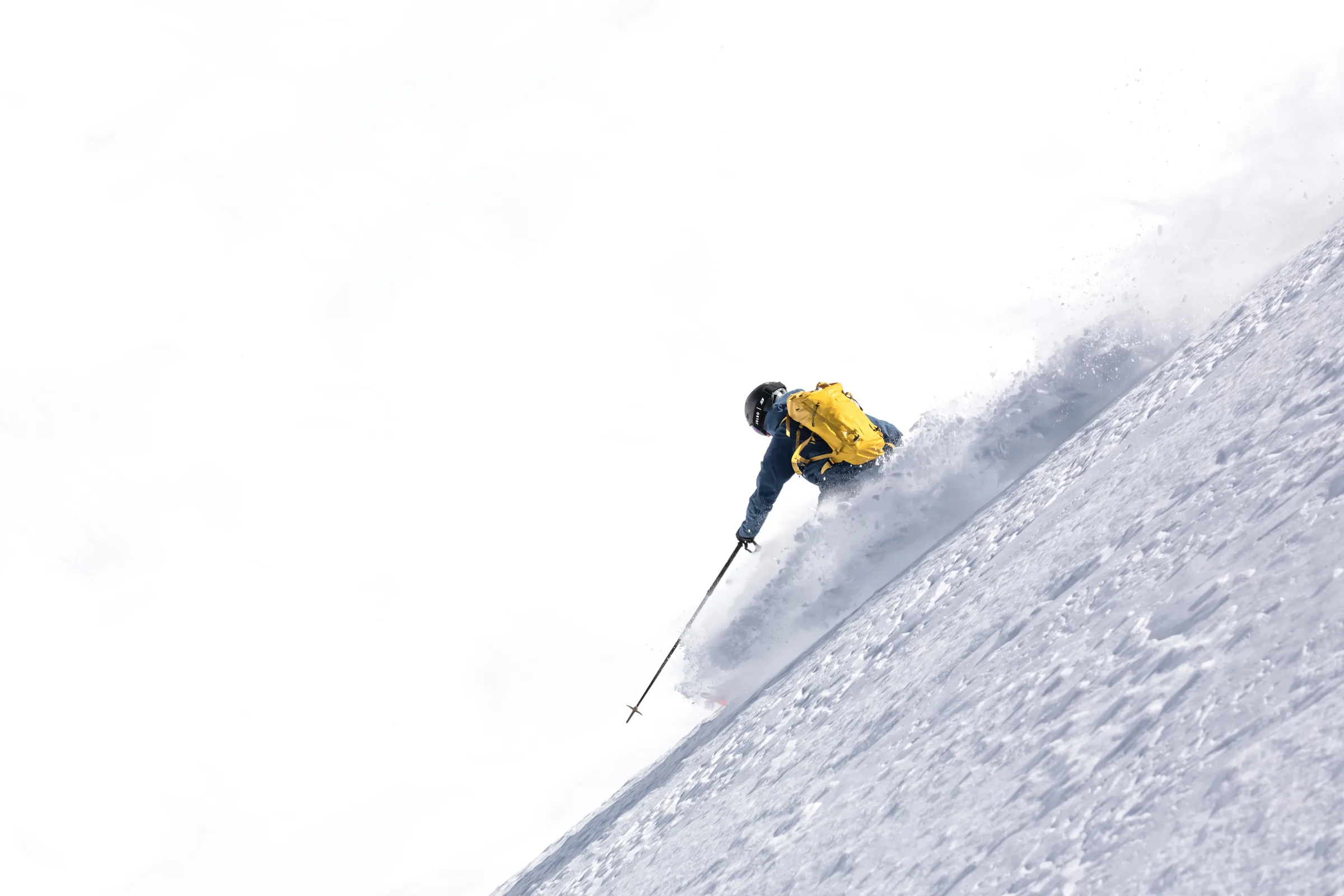 Seen from behind, Valentin Rapp skis down a steep snowy slope with a yellow backpack, reaching a pole downhill as snow sprays behind him.