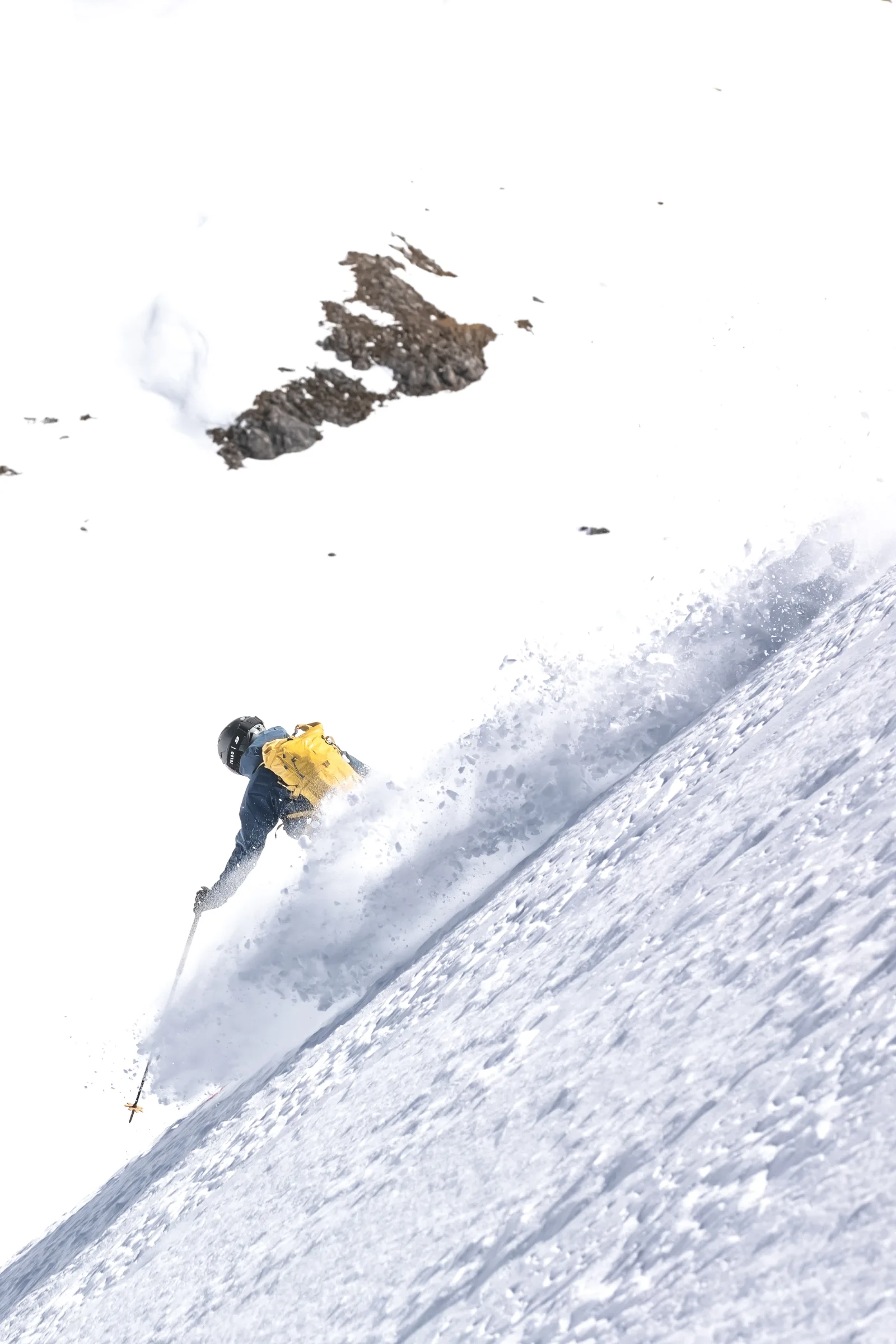 Valentin Rapp turns through deep snow on a steep slope below exposed rocks, his yellow backpack above a broad plume of powder.