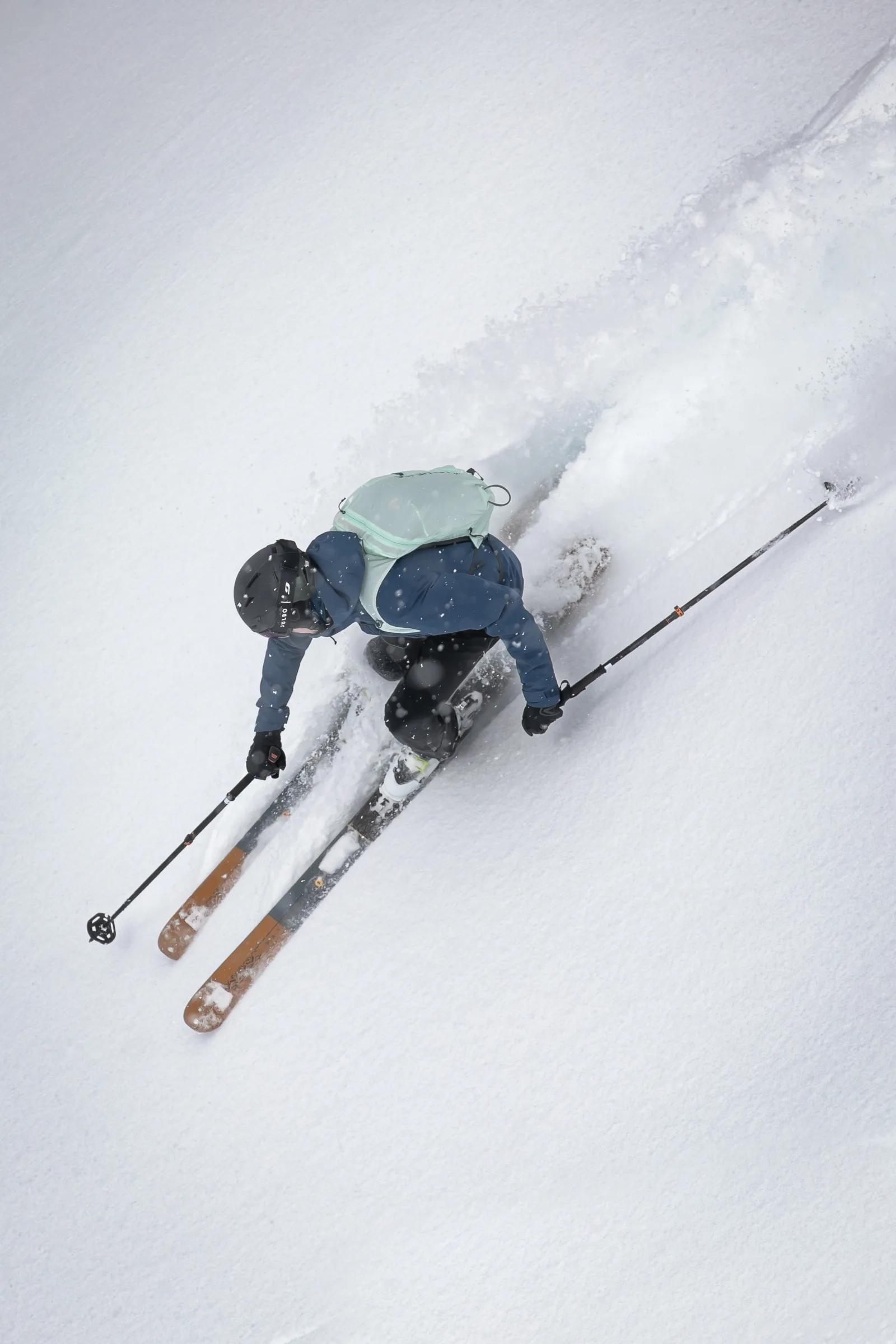 Seen from above, a skier in a blue jacket and pale green backpack crouches low through powder with ski poles extended to either side.