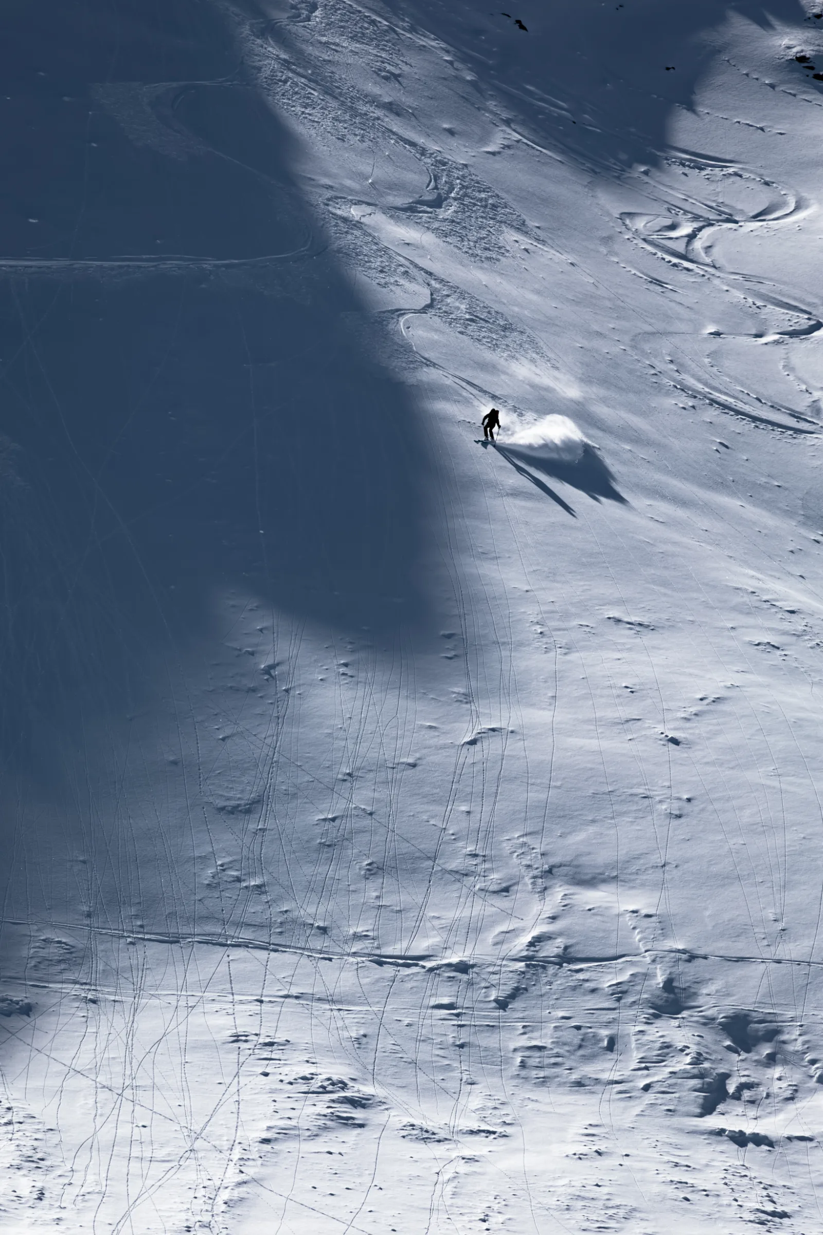 A lone skier turns across a broad, tracked snow slope, casting a long shadow beside a bright plume of powder.