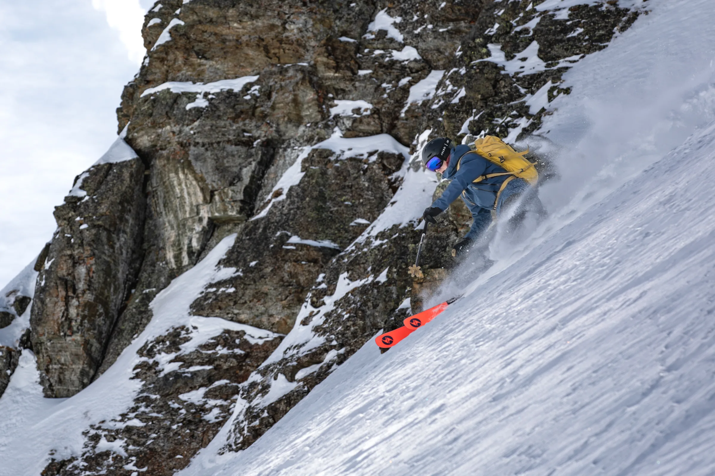Valentin Rapp skis down a steep snowy slope beside a rugged rock face, wearing a yellow backpack and riding bright red skis.