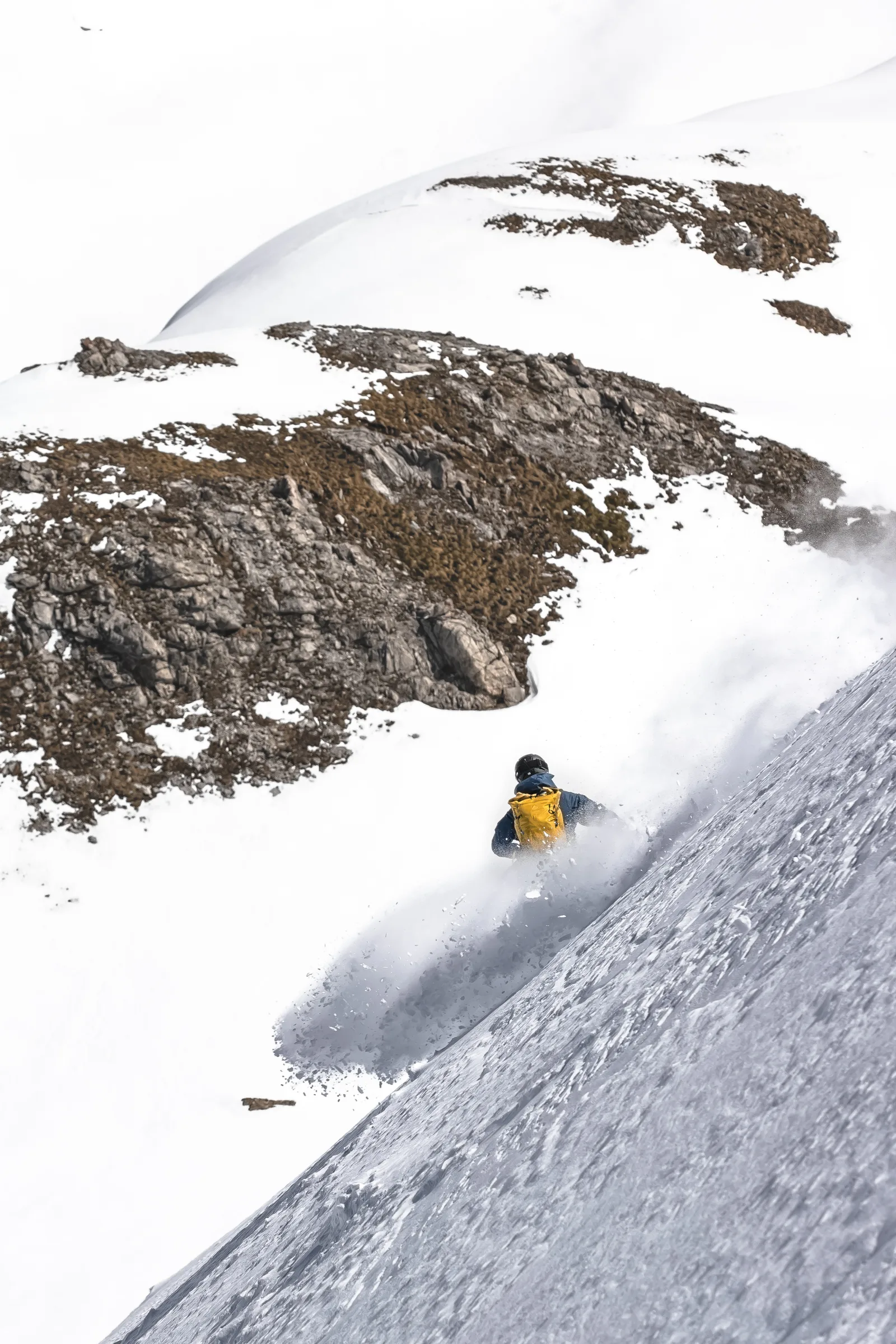 Seen from behind, Valentin Rapp skis through a large cloud of powder on a steep slope, with snow-covered rocky ridges beyond him.